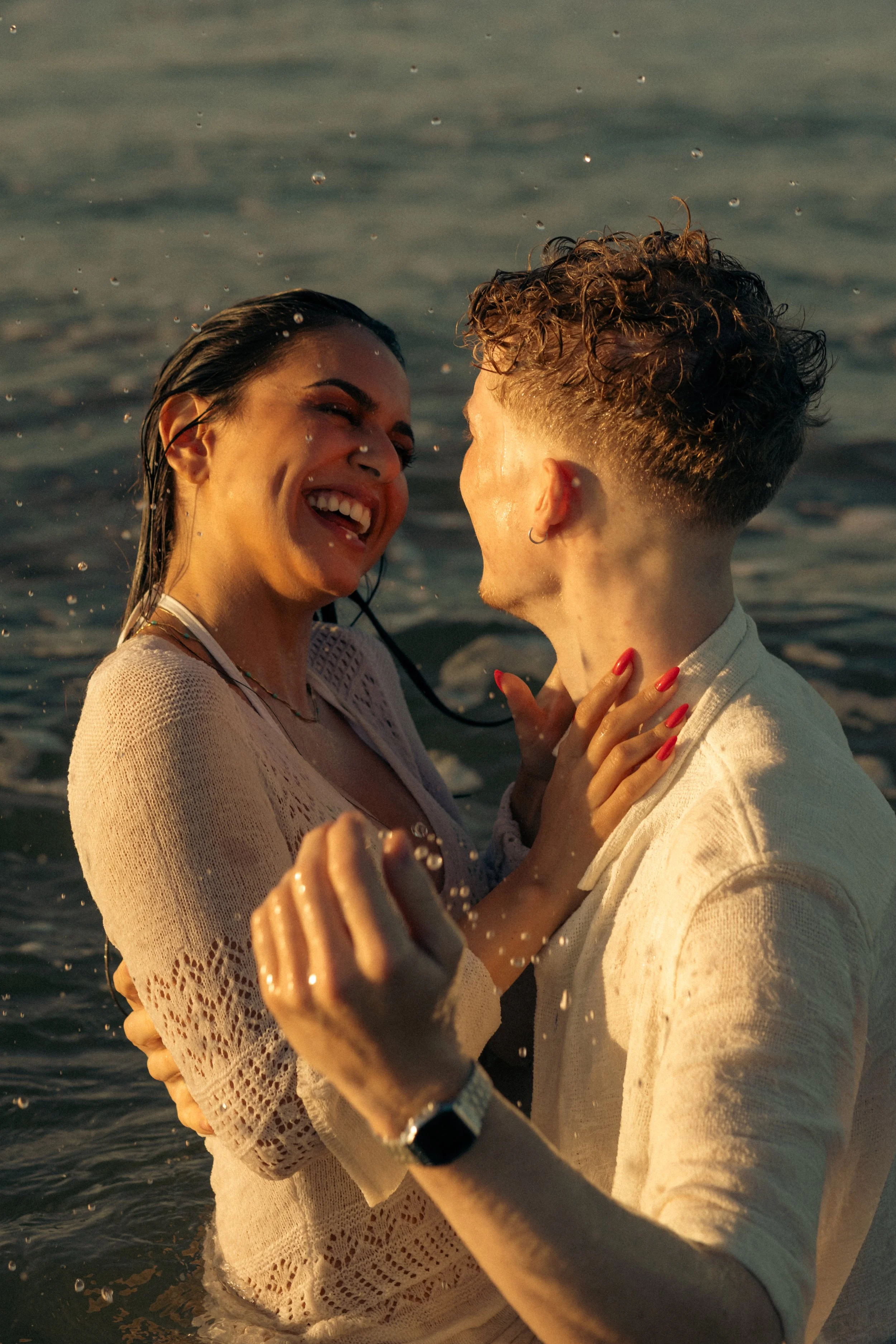 A joyful couple laughing and embracing in the water at the beach during sunset.