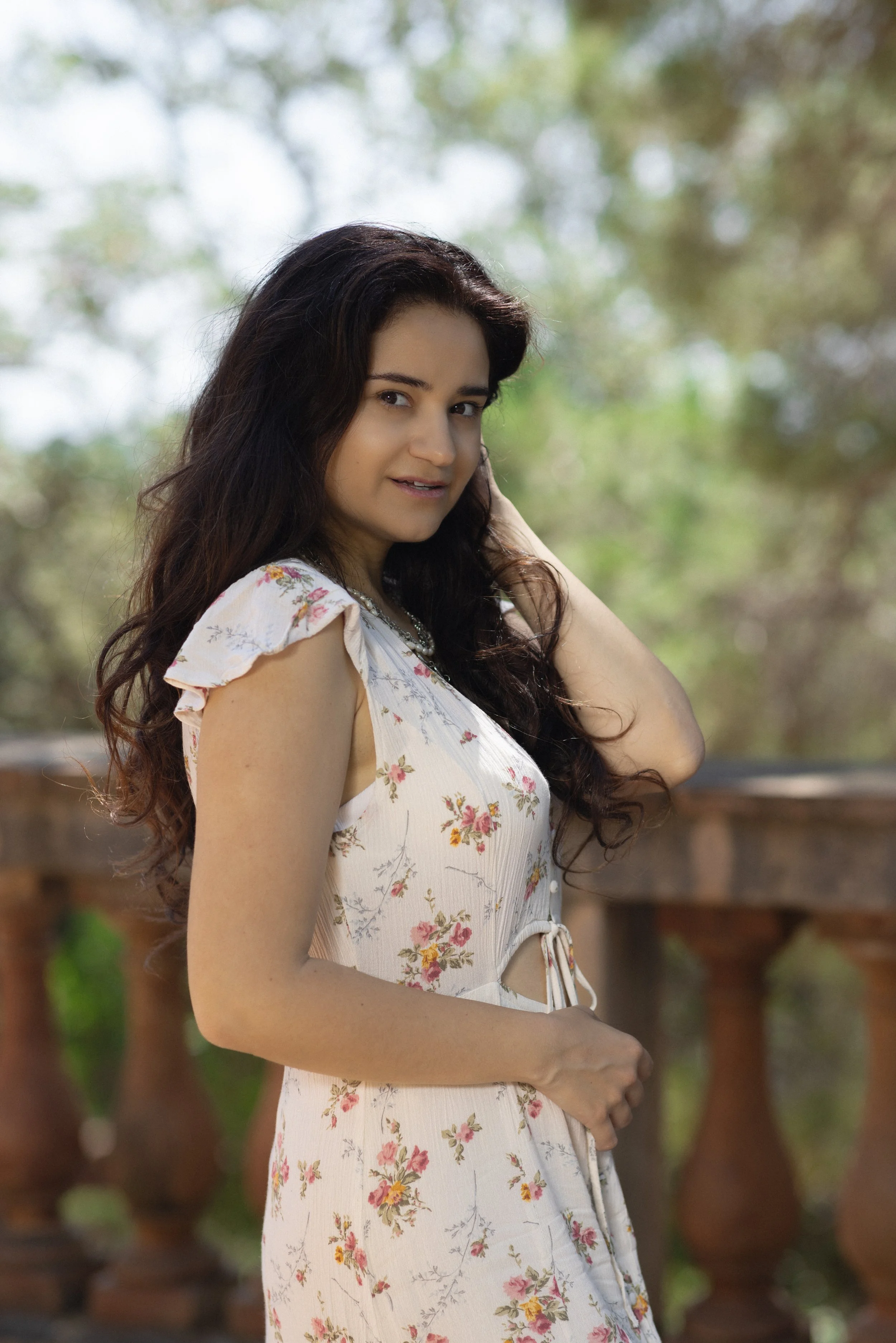 A woman with long dark hair wearing a white floral dress standing outdoors on a wooden balcony with blurred greenery in the background.