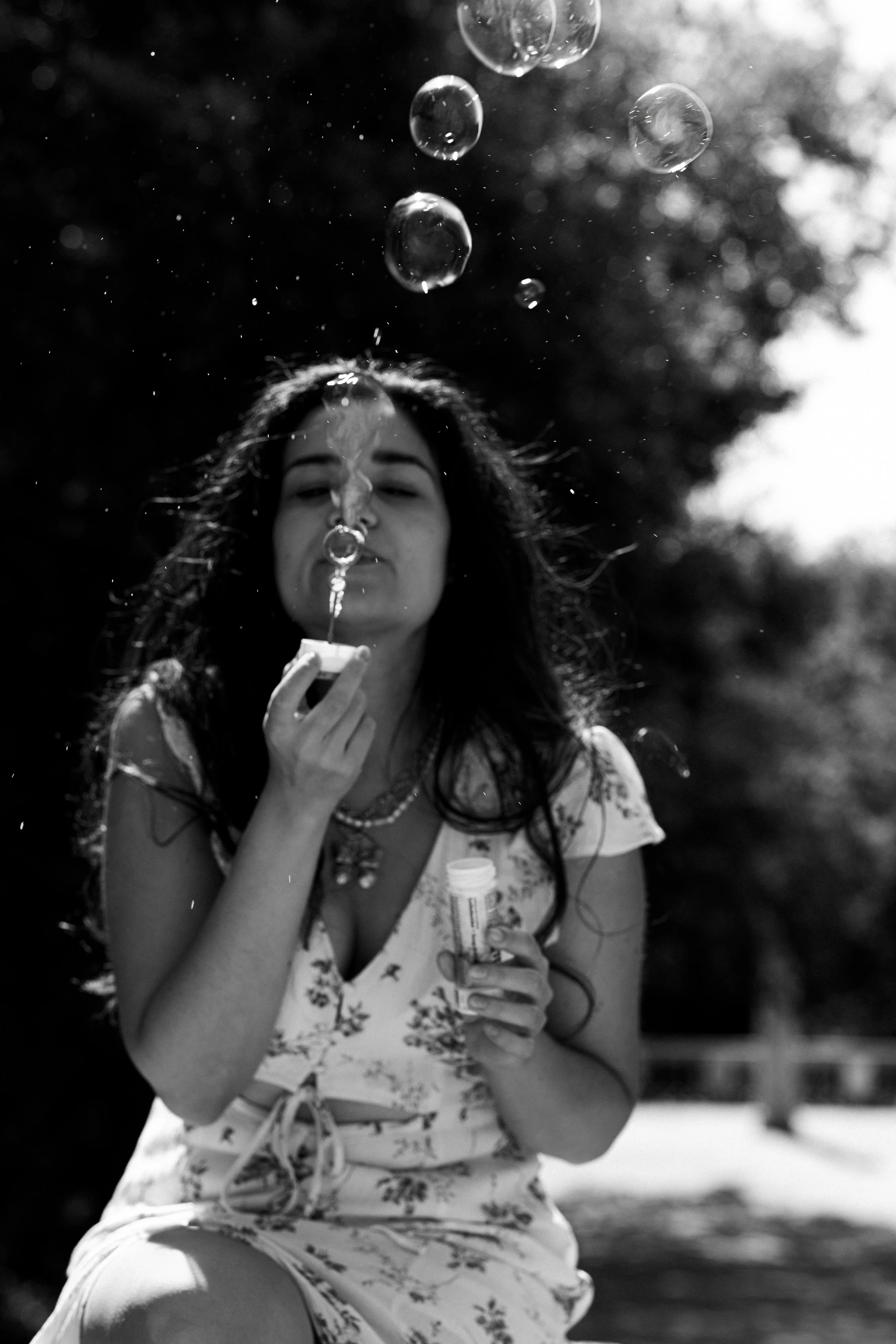 A woman blowing bubbles outdoors, holding a bubble wand and container, with sunlight backlighting her, in a black and white photo.
