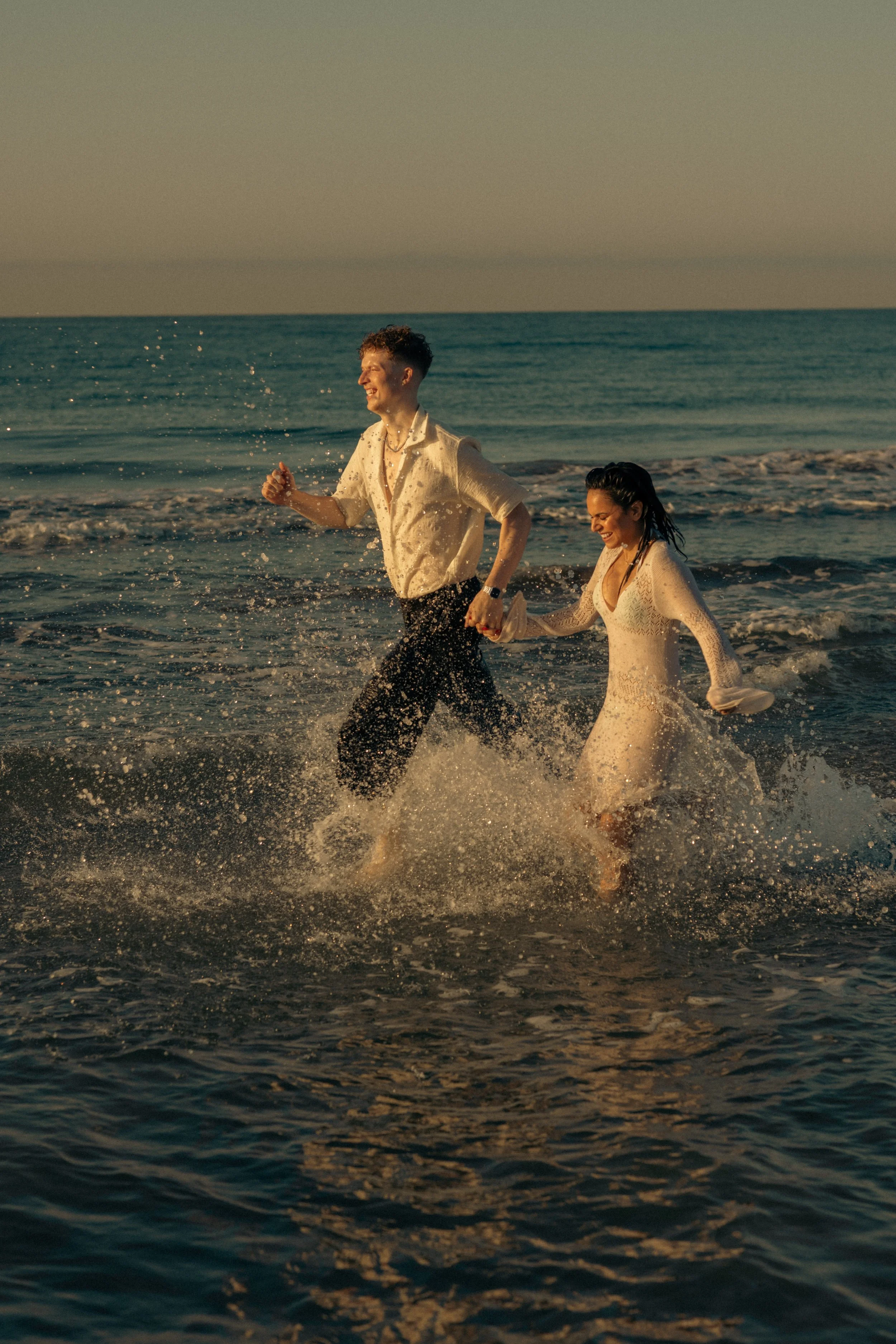 A happy couple in wedding attire running through the ocean waves at sunset.