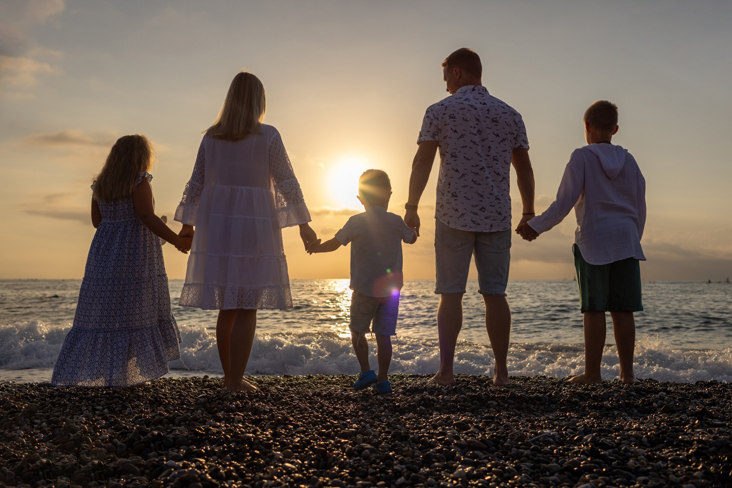 Family holding hands and facing the sunset on a beach with pebbles.