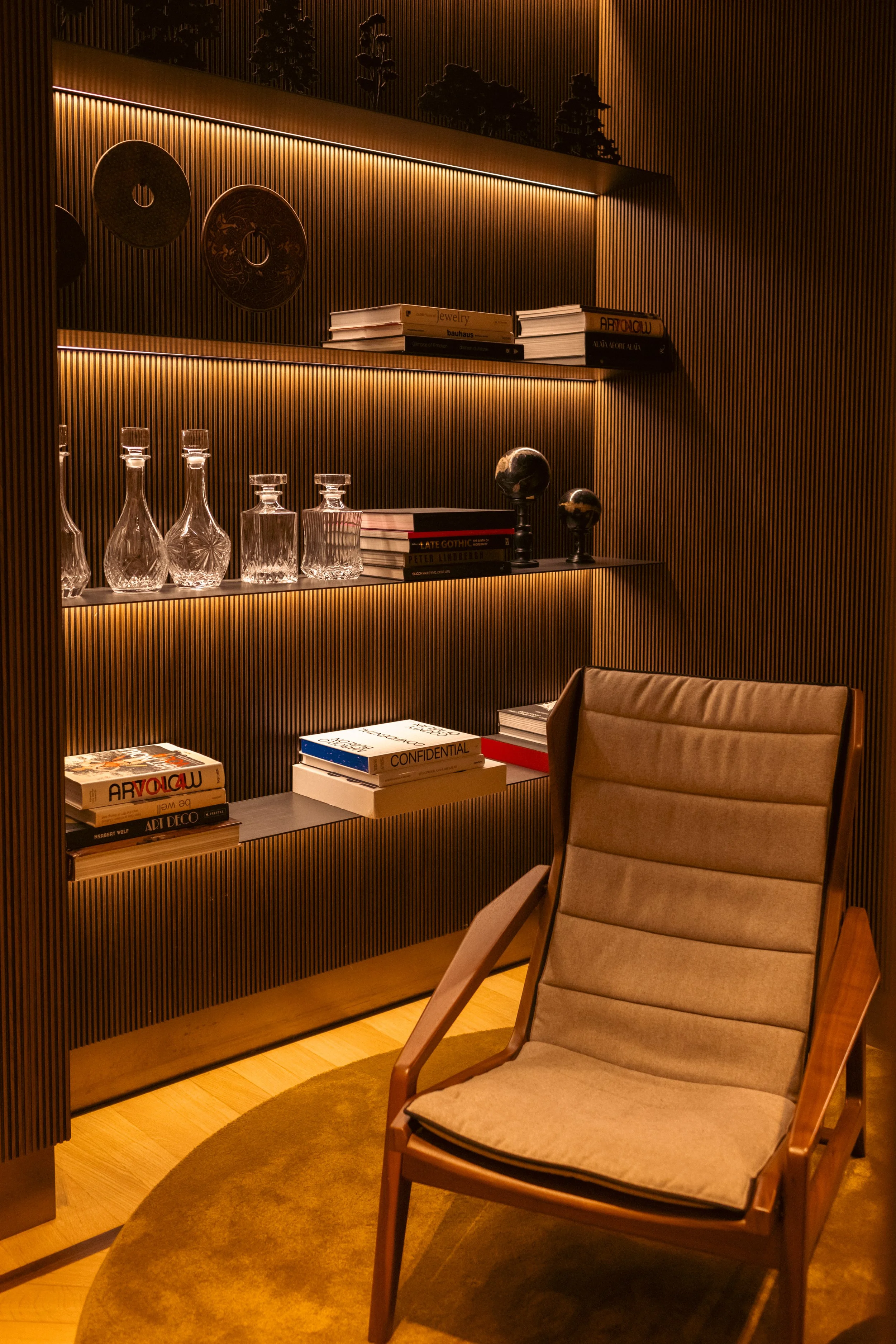A cozy corner with a beige chair in front of illuminated wooden shelves holding books, glass decanters, and decorative globes.
