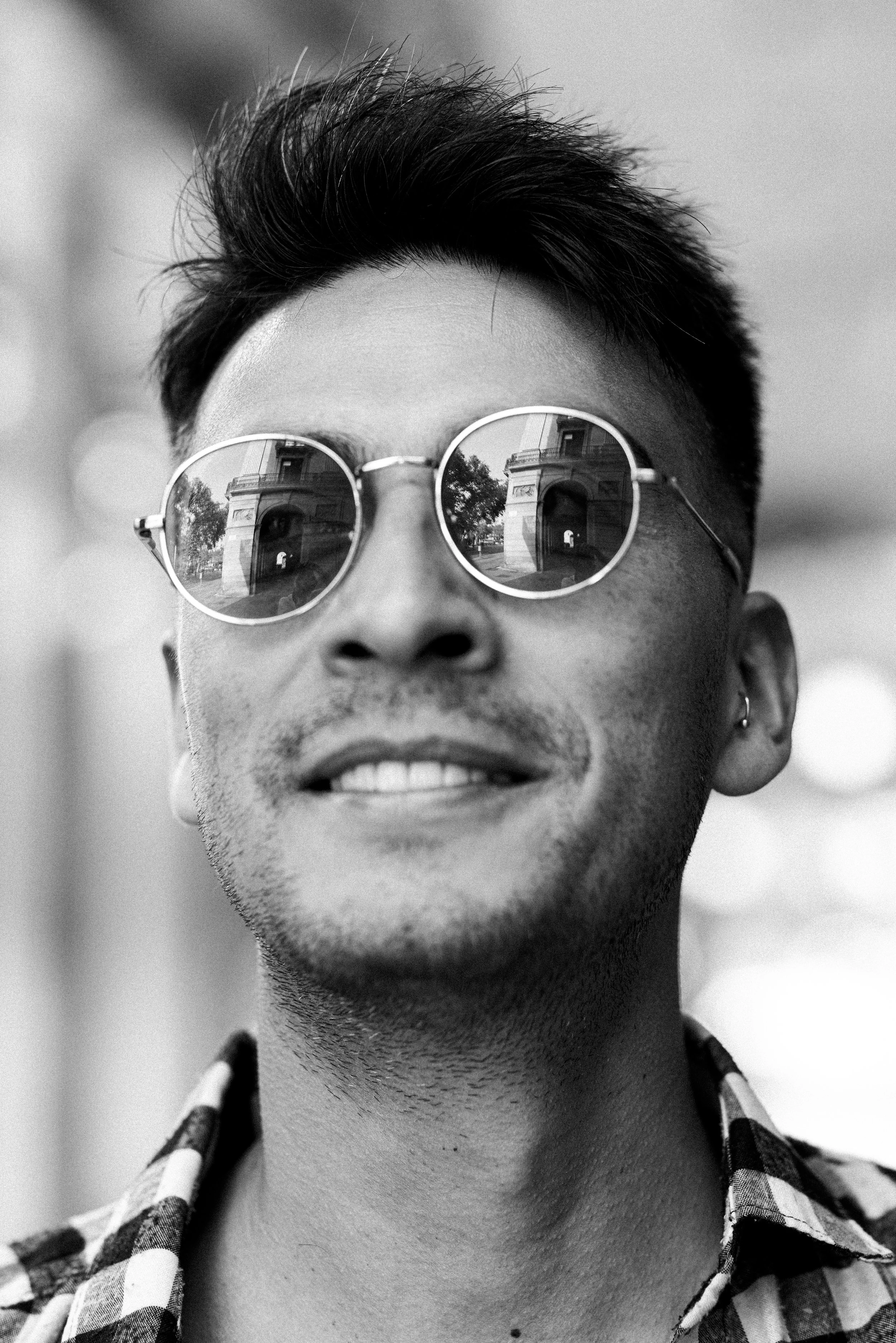 Black and white close-up photo of a young man with sunglasses reflecting buildings, smiling, wearing a plaid shirt.