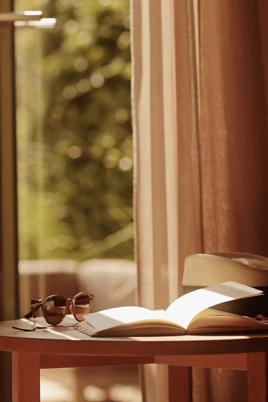 A pair of sunglasses and an open book on a wooden table near a window with sunlight shining through curtains.
