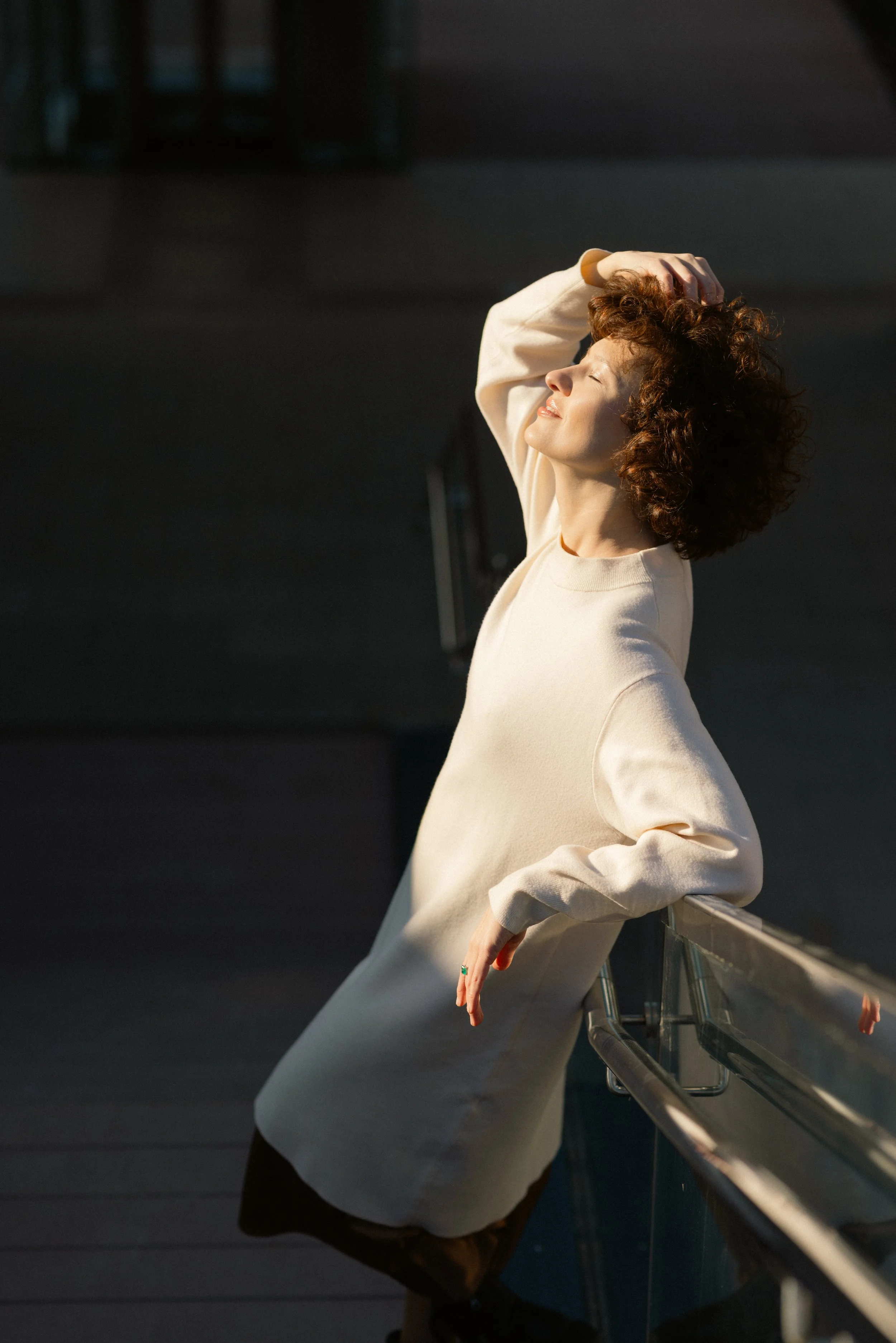 A woman with curly hair wearing a cream-colored coat, basking in sunlight with her eyes closed, leaning against a railing on a balcony or terrace.