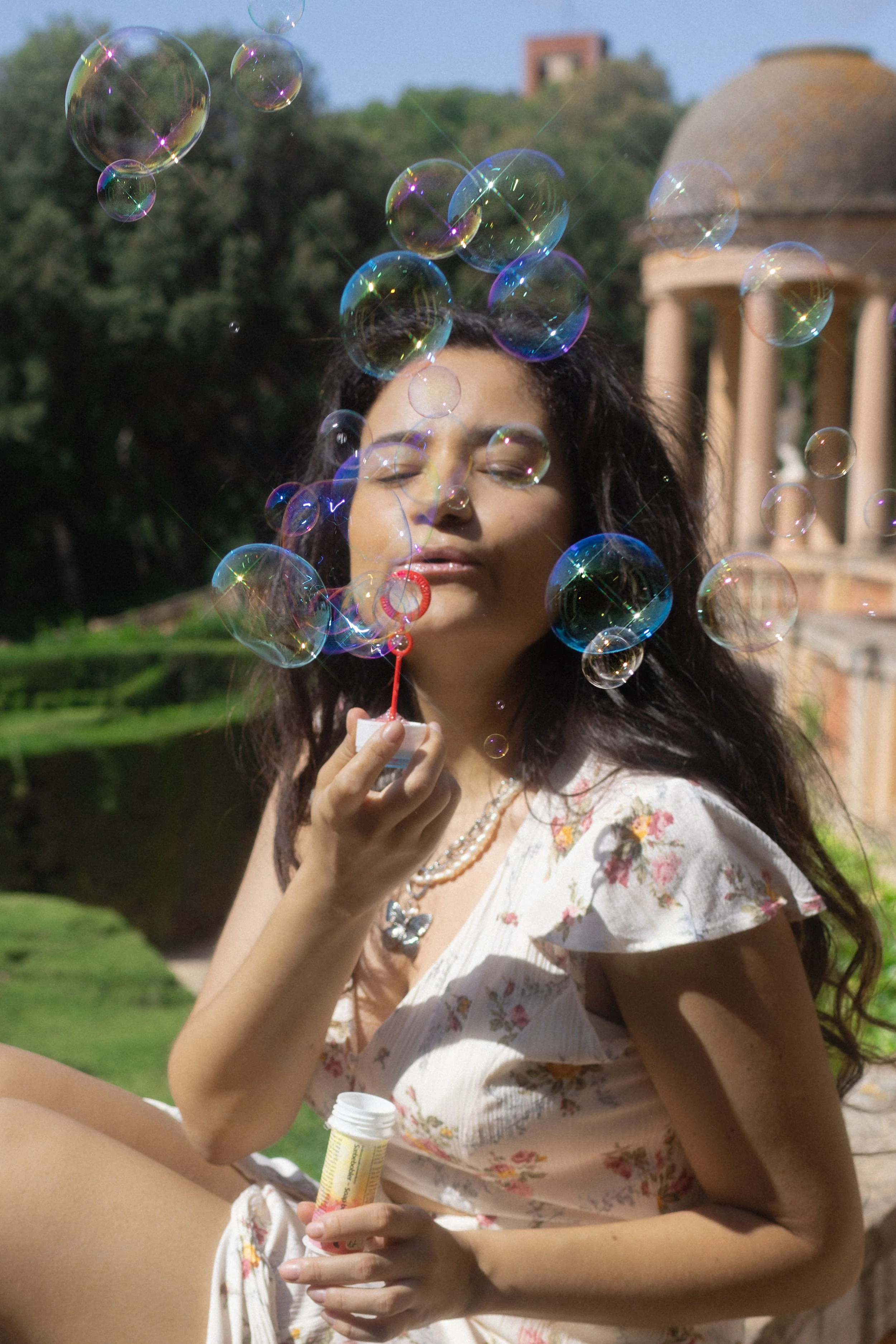 A woman with long dark hair blowing bubbles outdoors on a sunny day, with a classical pavilion and lush greenery in the background.