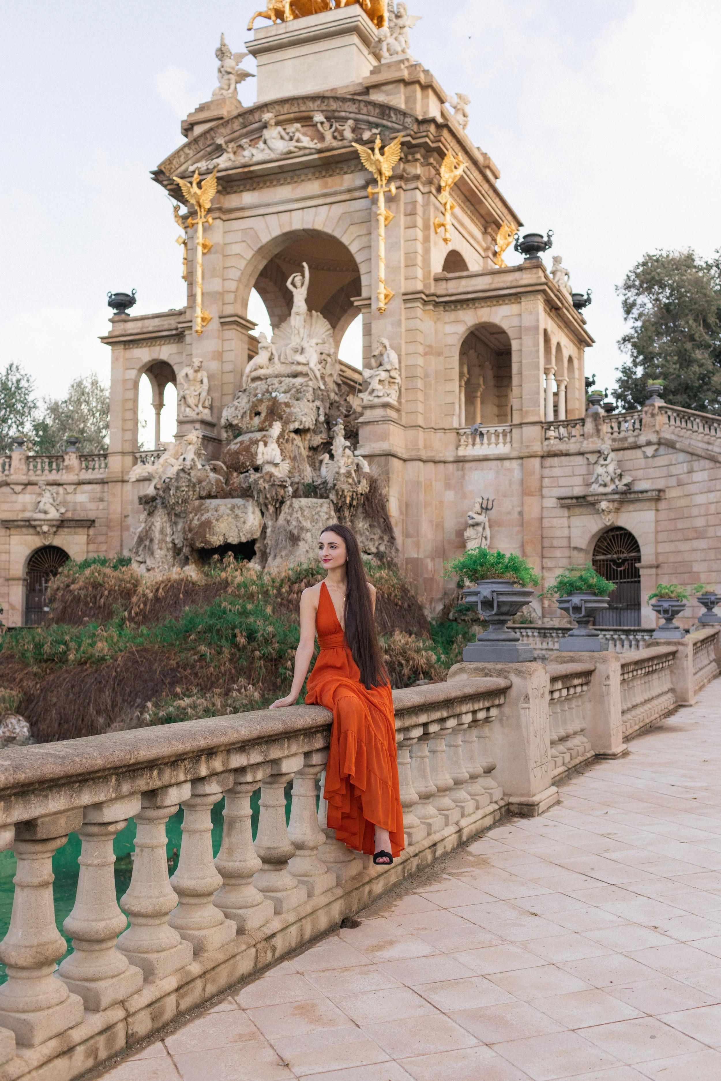 A woman in an orange dress sitting on a stone balustrade in front of an ornate, historic fountain and architectural structure with sculptures and statues, during daylight.