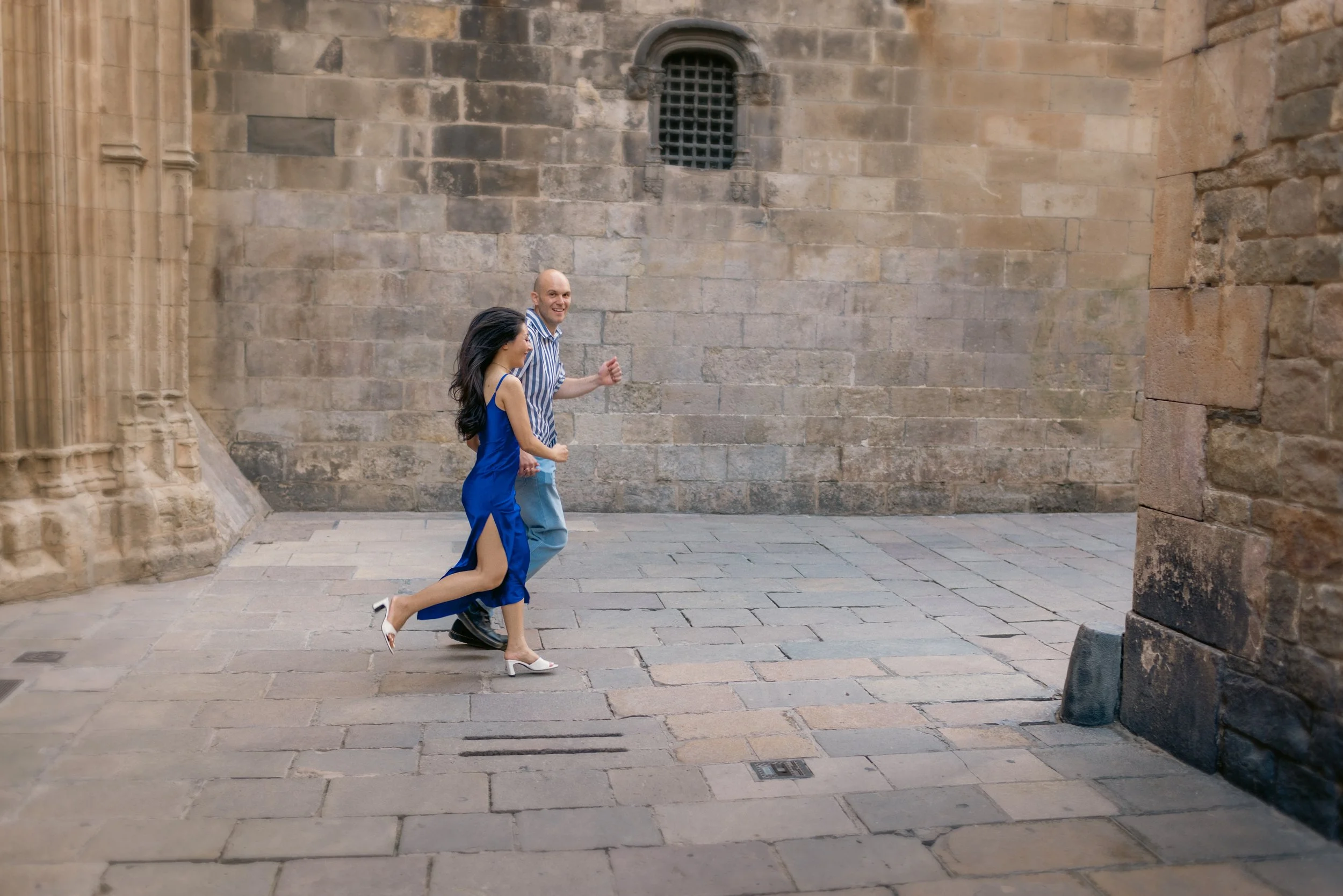 A man and woman walking and smiling along a stone-paved street near a historic stone building, with the woman wearing a blue dress with thigh-high slit and white heels, and the man dressed in a striped shirt and gray pants.