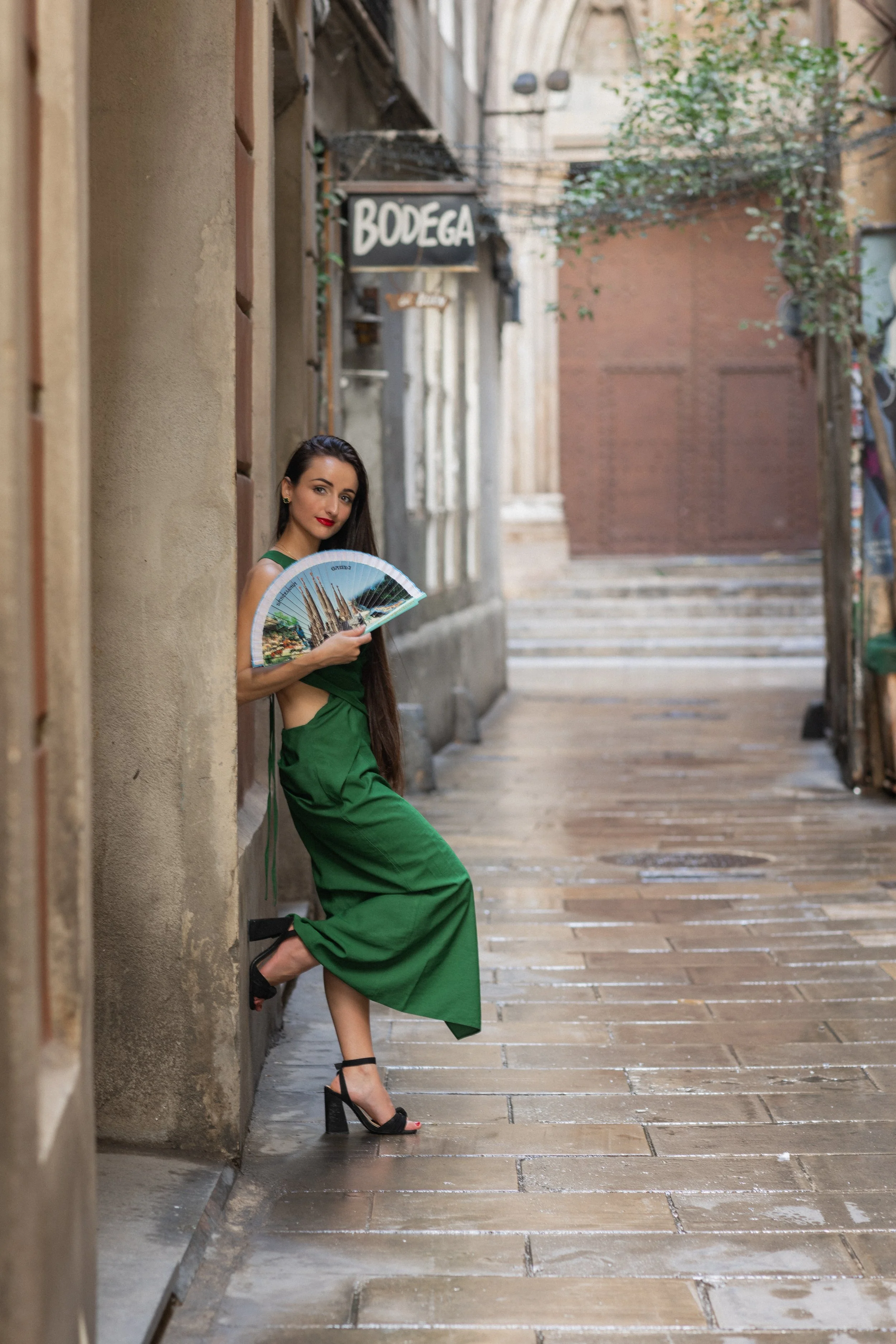 A woman with long dark hair, wearing a green dress and black high heels, stands in a narrow alleyway holding a decorative fan with a cityscape image, looking at the camera.