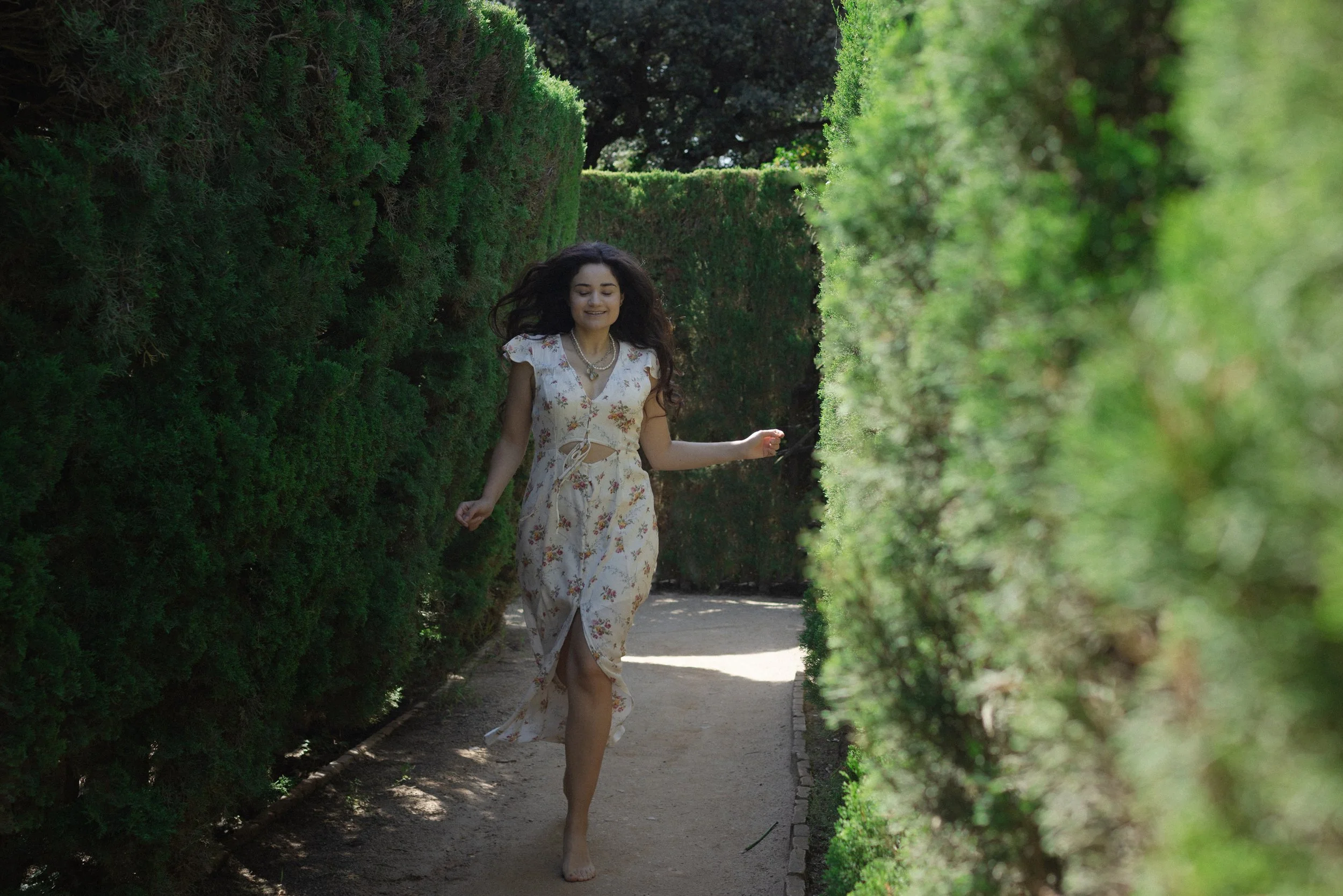 A young woman with long dark hair, wearing a white floral dress, walking barefoot along a narrow garden pathway between green hedges, smiling and enjoying the outdoors.