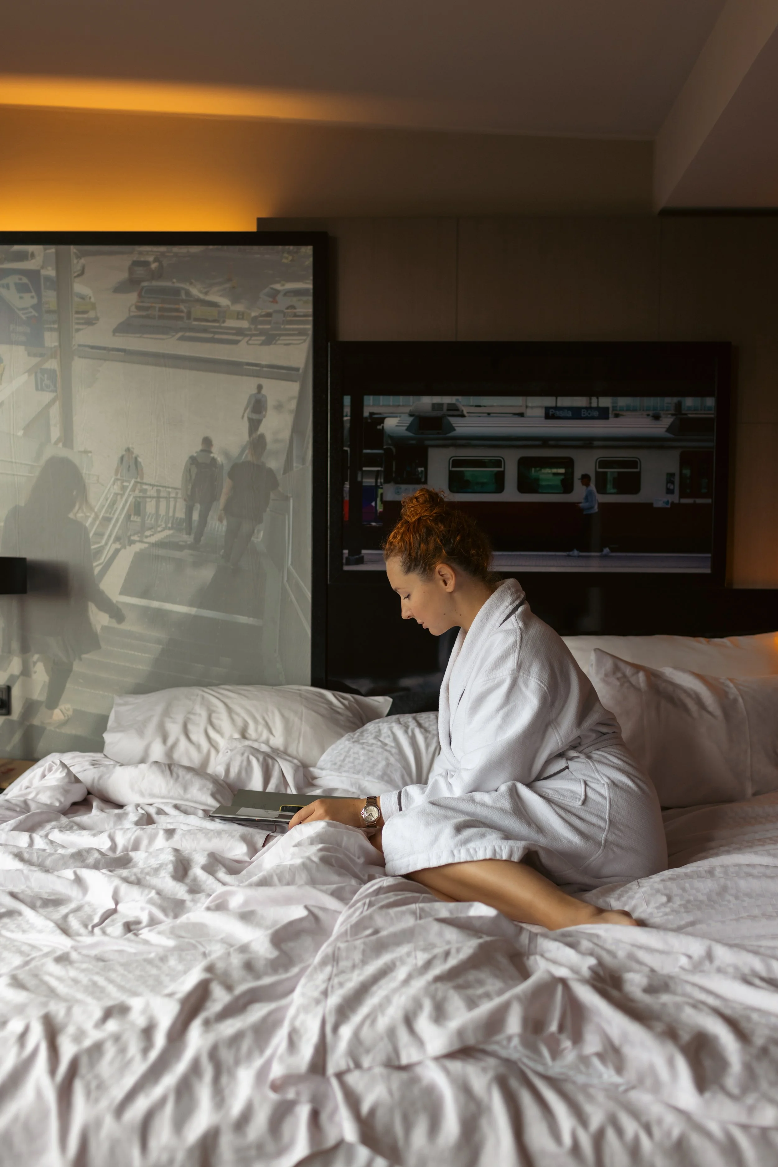 A woman in a white hotel bathrobe sitting on a bed, looking at a tablet, in a hotel room with windows showing passing trains and people walking on stairs outside.