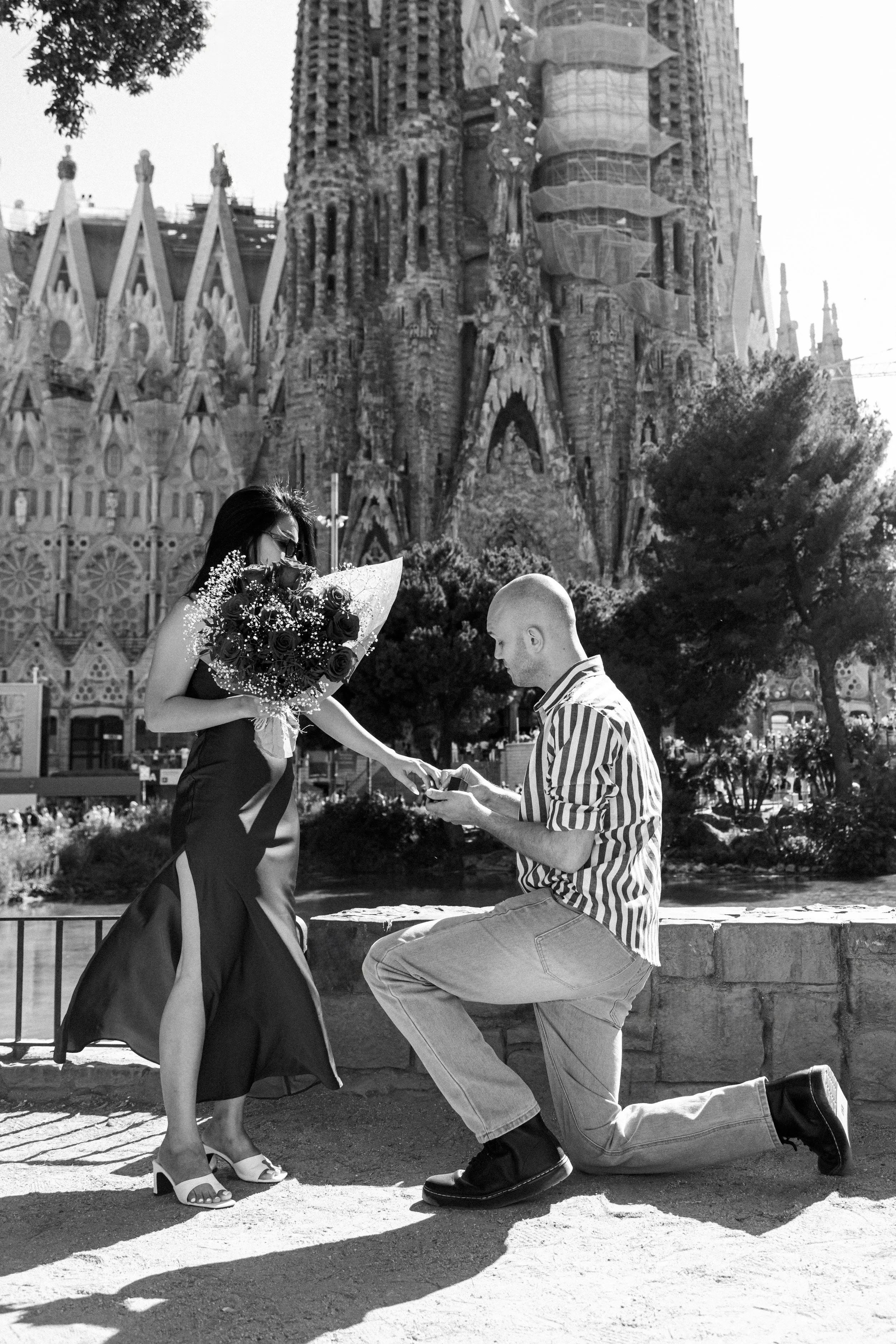 A man kneeling on one knee presenting an open ring box to a woman holding a bouquet of flowers, in front of the Sagrada Familia in Barcelona, Spain.
