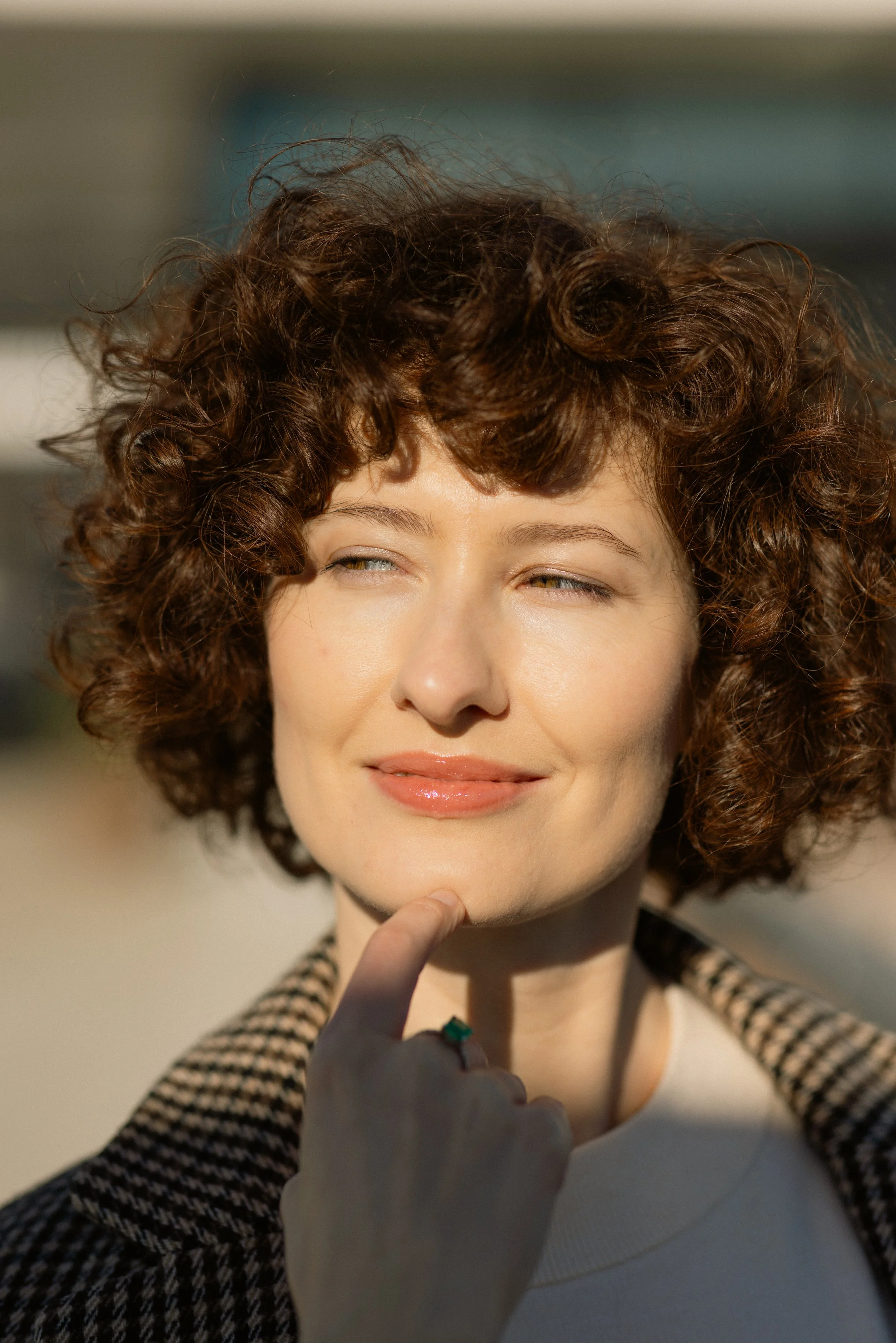A woman with curly brown hair smiling and touching her chin with her hand, outdoors in sunlight.