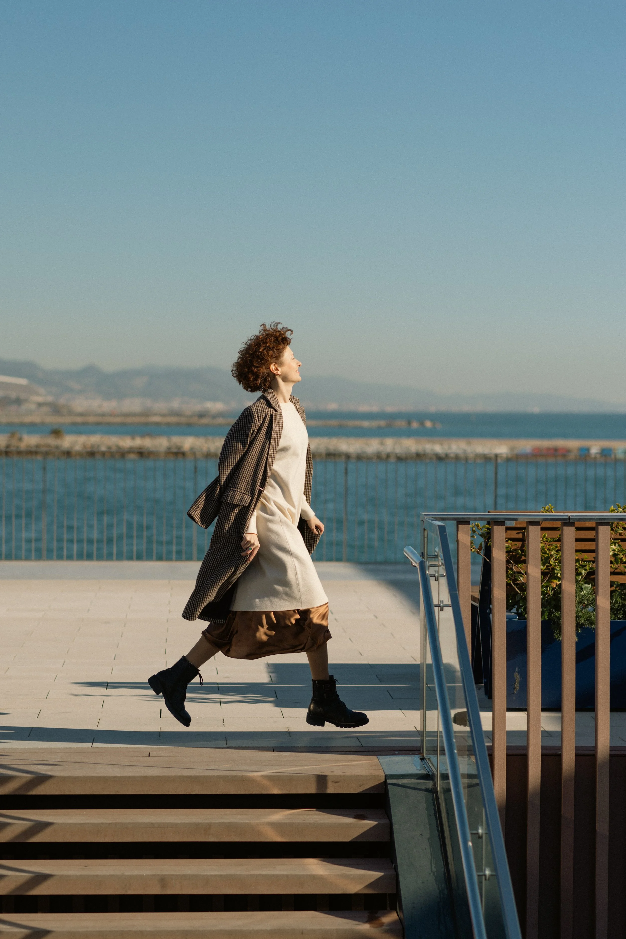 Woman walking outdoors near water with mountains in the background, wearing a long coat, dress, and combat boots.
