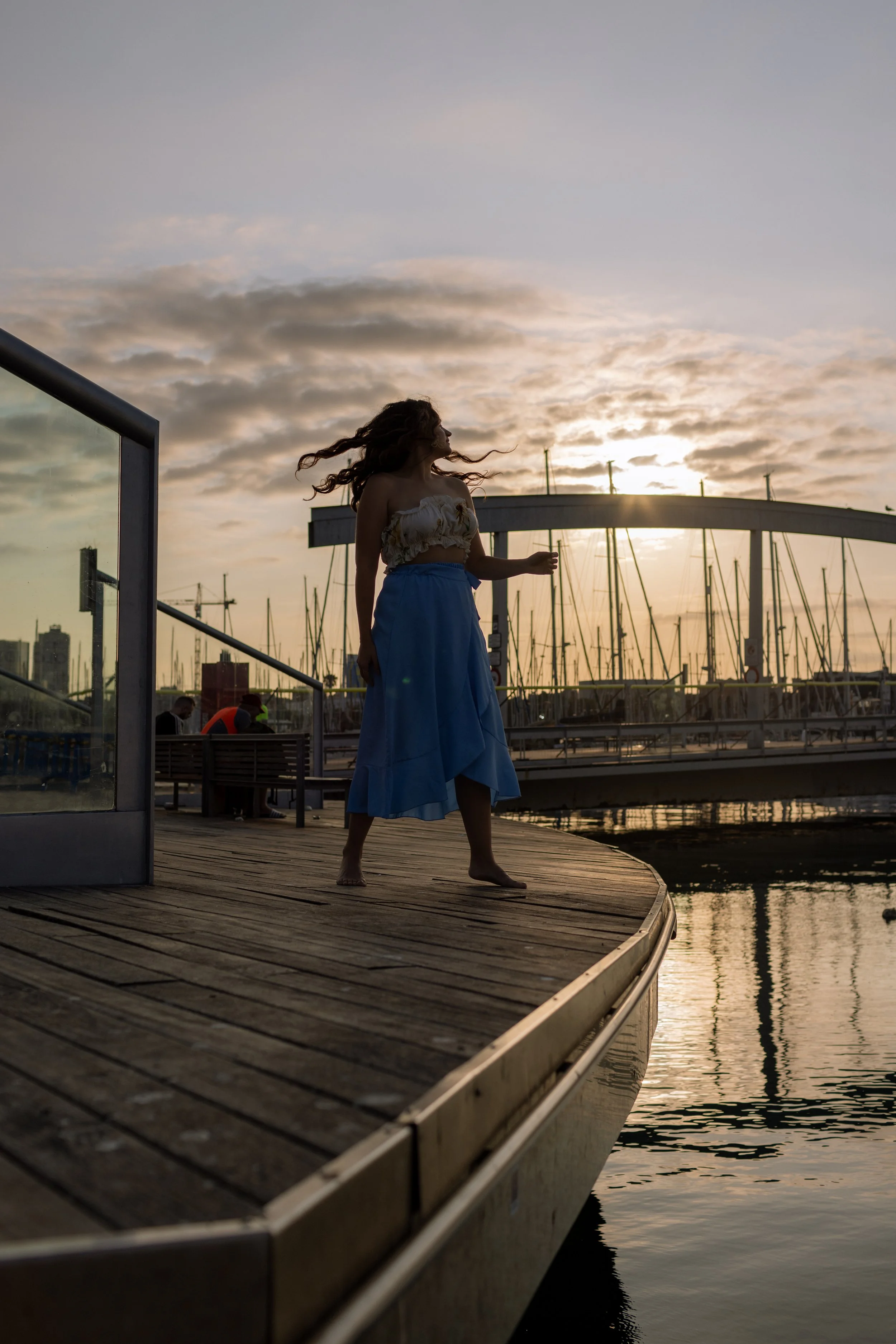 A woman in a white top and blue skirt stands on a wooden dock by the water at sunset, with sailboats and a bridge in the background.
