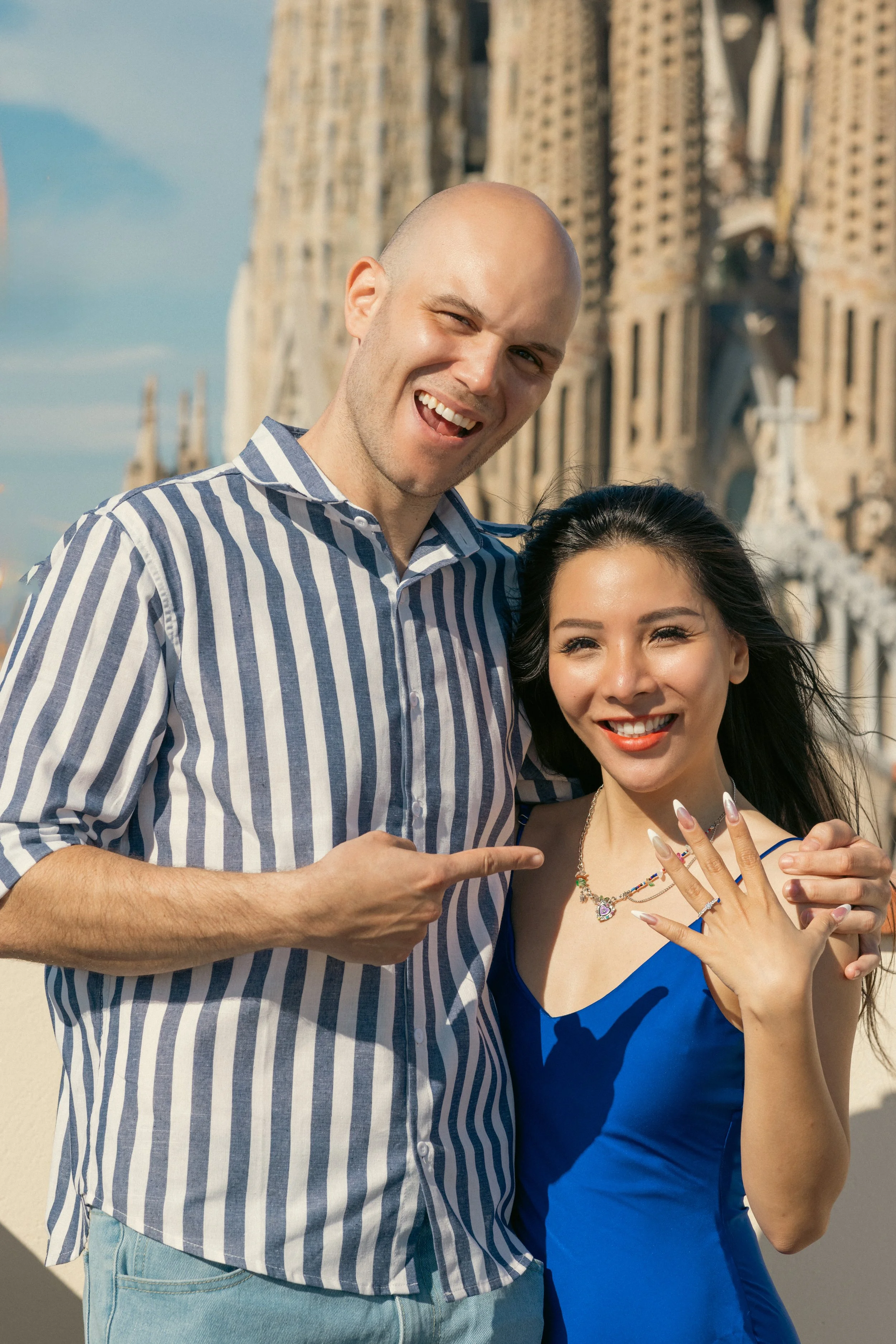 A smiling man wearing a striped shirt points at a woman in a blue dress with jewelry, showing engagement ring standing near a historic church with a cloudy sky in the background.