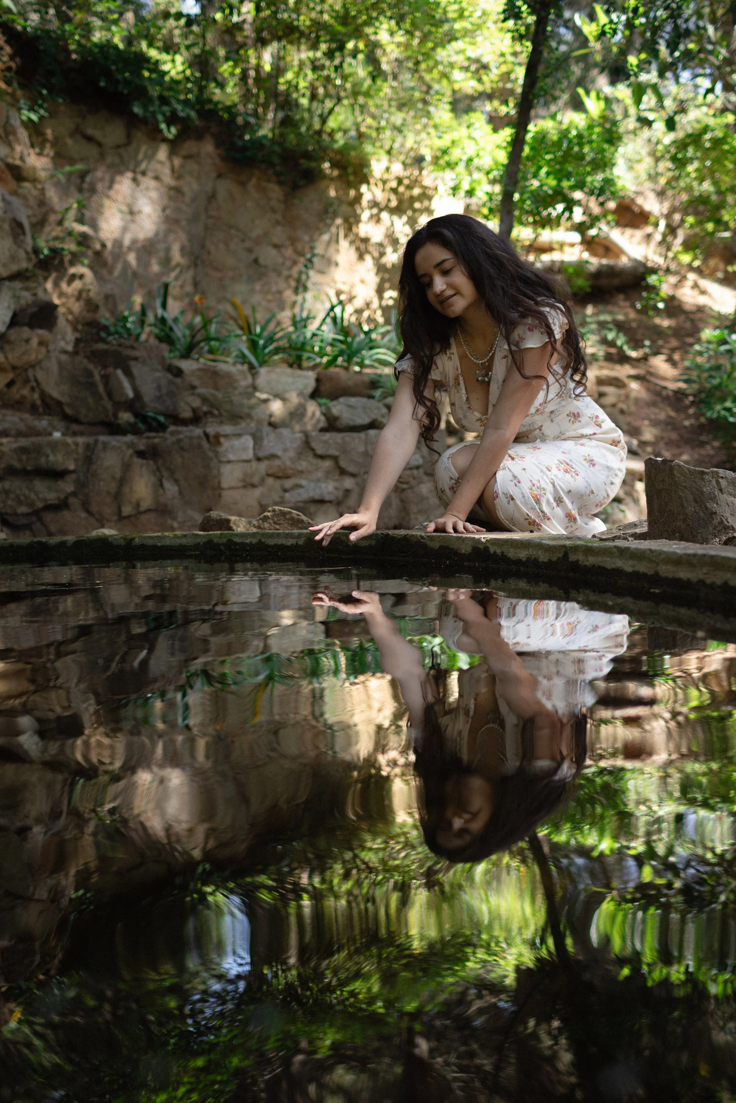 A woman in a floral dress crouches by a water feature in a garden, touching the water with her hand, with her reflection visible in the water.