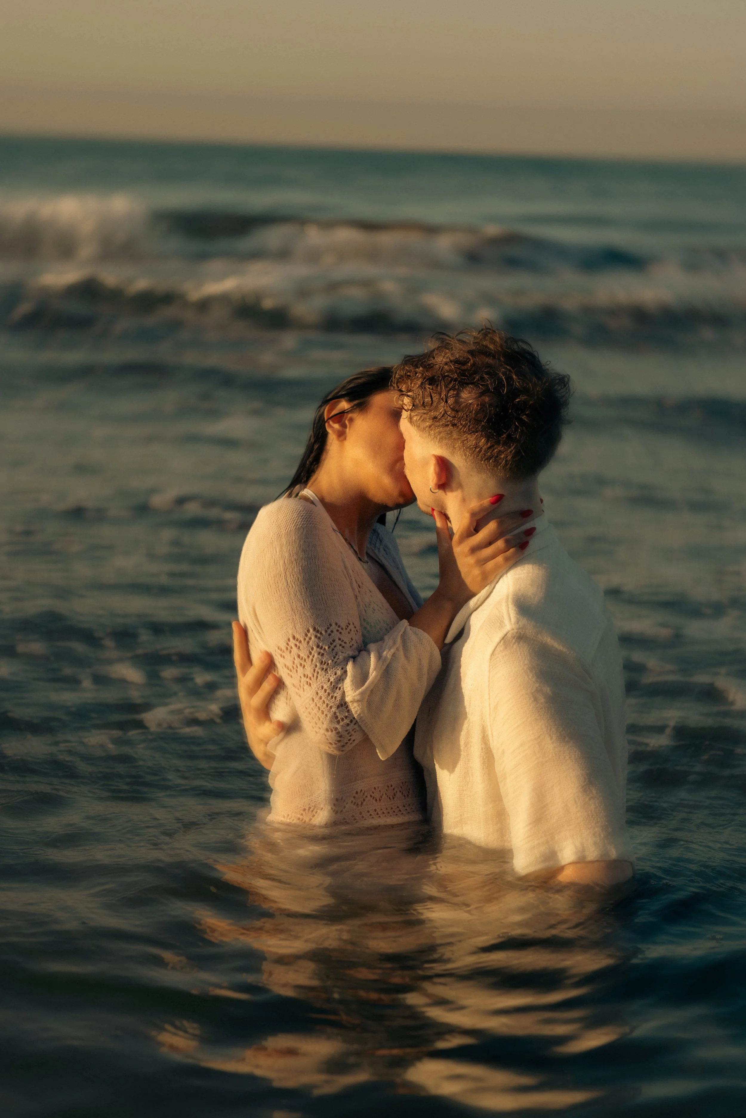 A couple kissing in the ocean during sunset, with waves in the background.