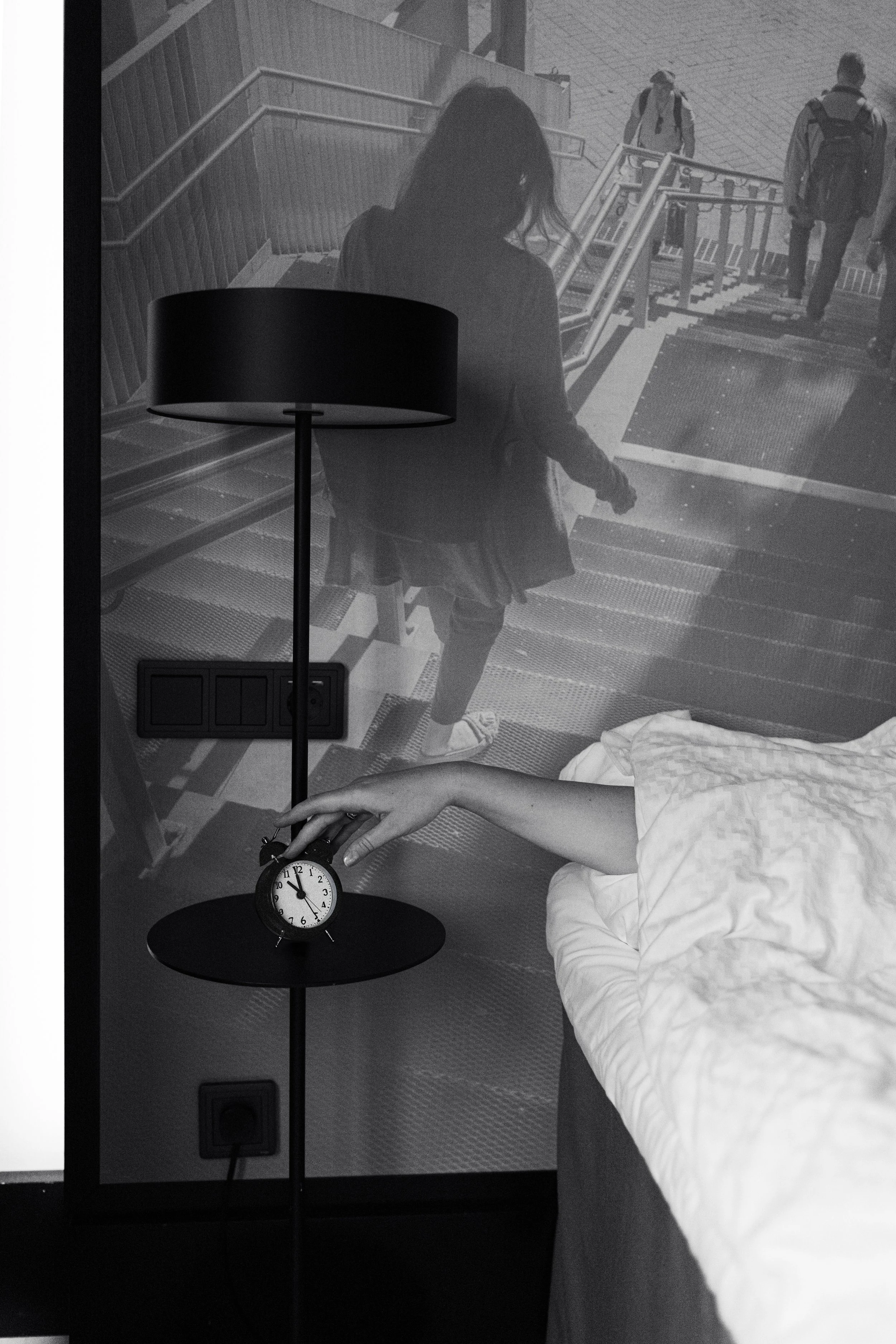 Black and white photo showing a person in bed reaching towards a small alarm clock on a bedside table, with a projection on the wall in front of the bed featuring a woman walking up stairs outside, viewed from behind.