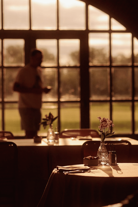 A dimly lit room with a table set for dining, decorated with a flower vase, and a man standing in the background near large windows showing an outdoor scene at sunset.
