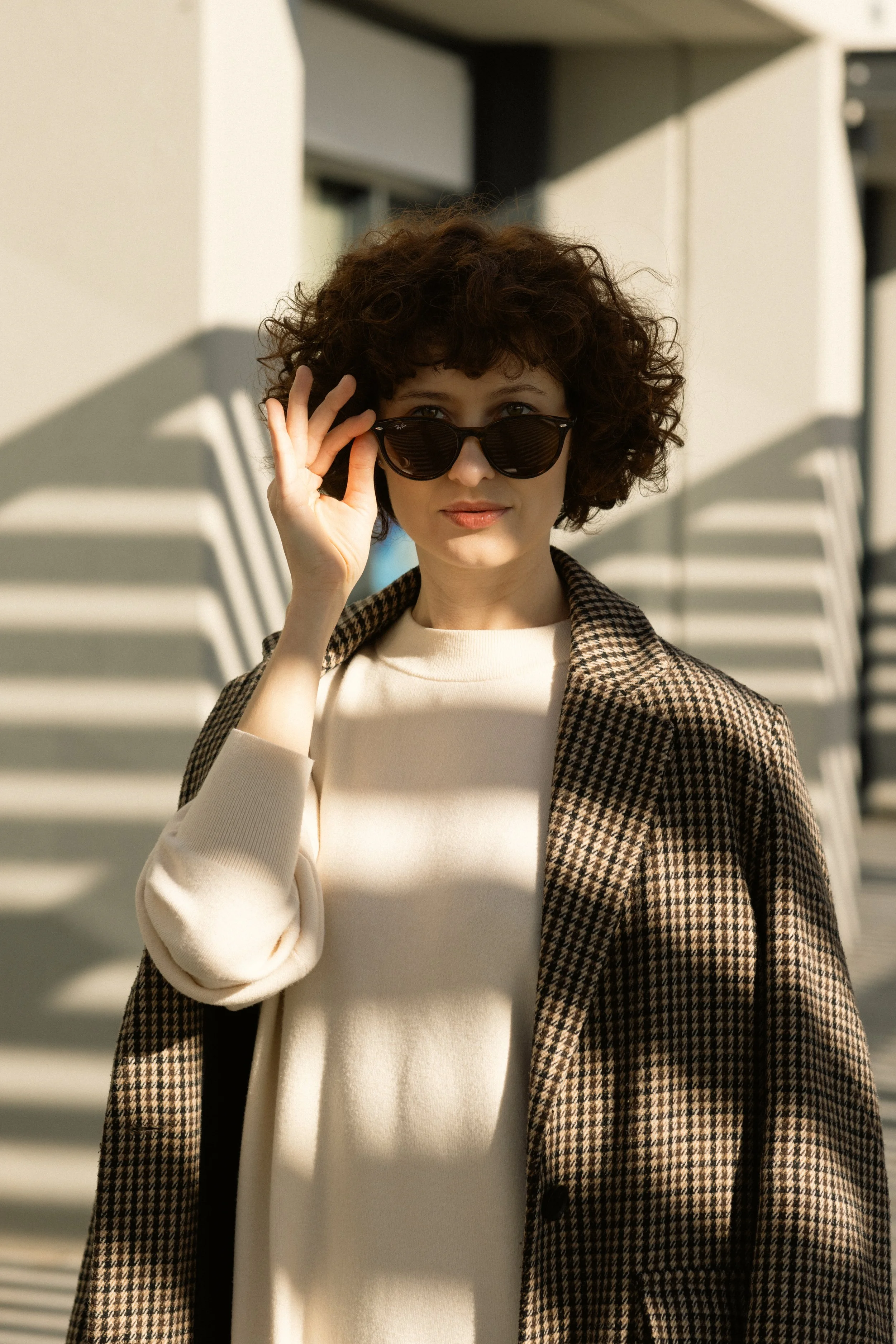 Woman with curly hair wearing black sunglasses, a cream sweater, and a checkered blazer outdoors with shadows cast by architectural elements.