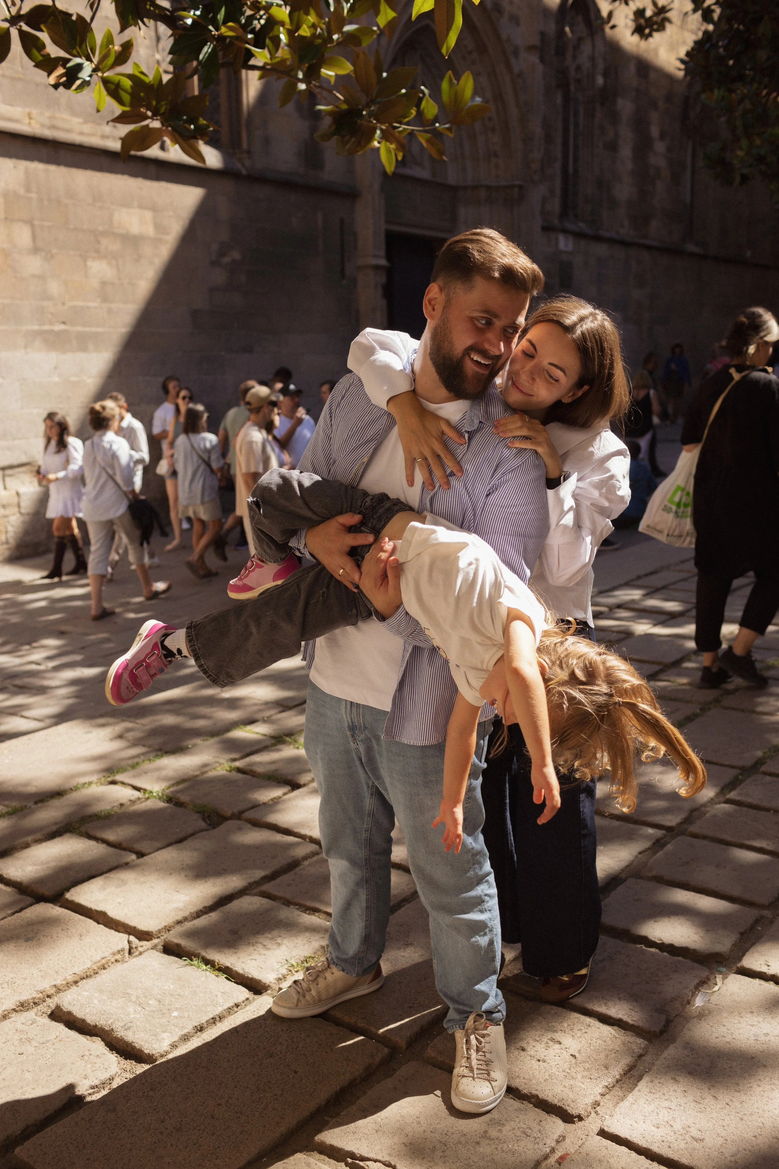 A happy family of four outside on a sunny day, with the father carrying his young daughter and the mother holding their older daughter.
