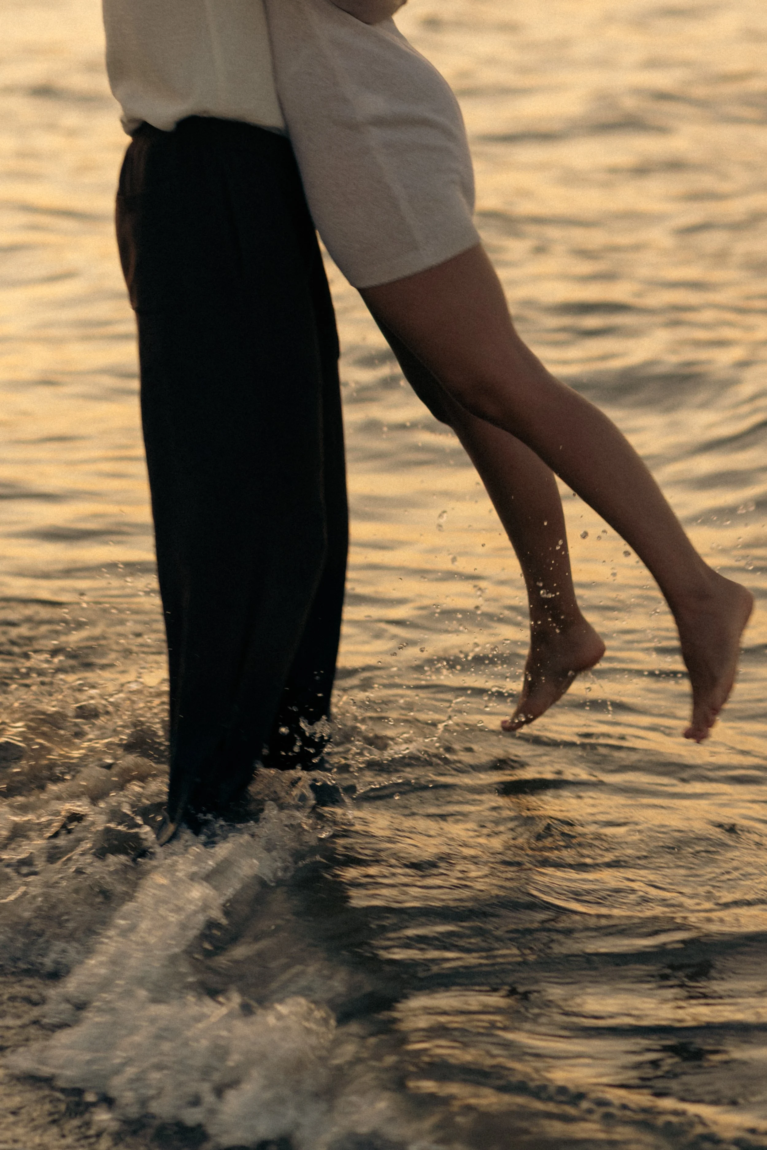 A person wearing a white shirt and dark pants standing in shallow water, holding another person who is mid-air, over the water, during sunset.
