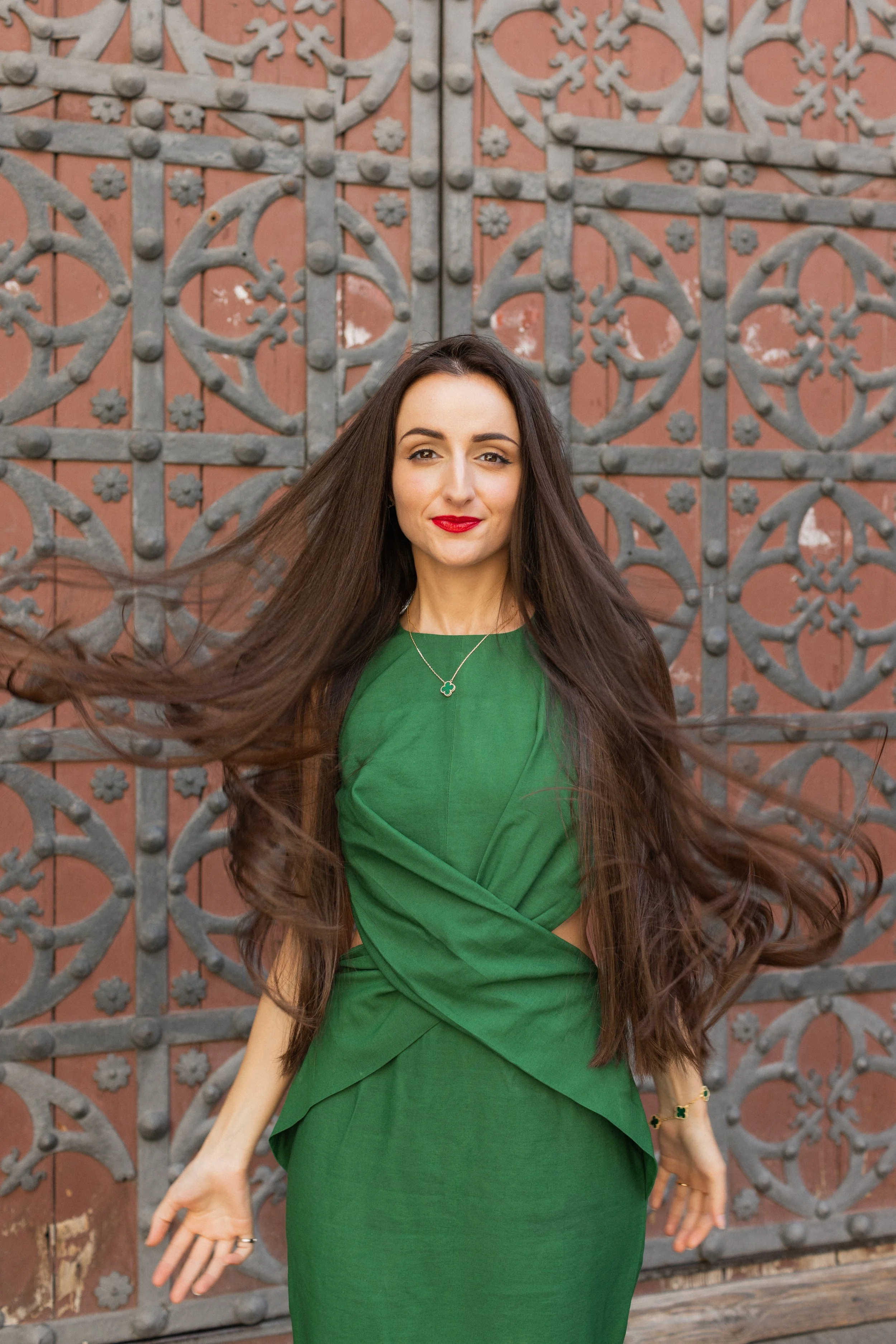A woman with long dark hair wearing a green dress poses in front of an ornate metal gate.