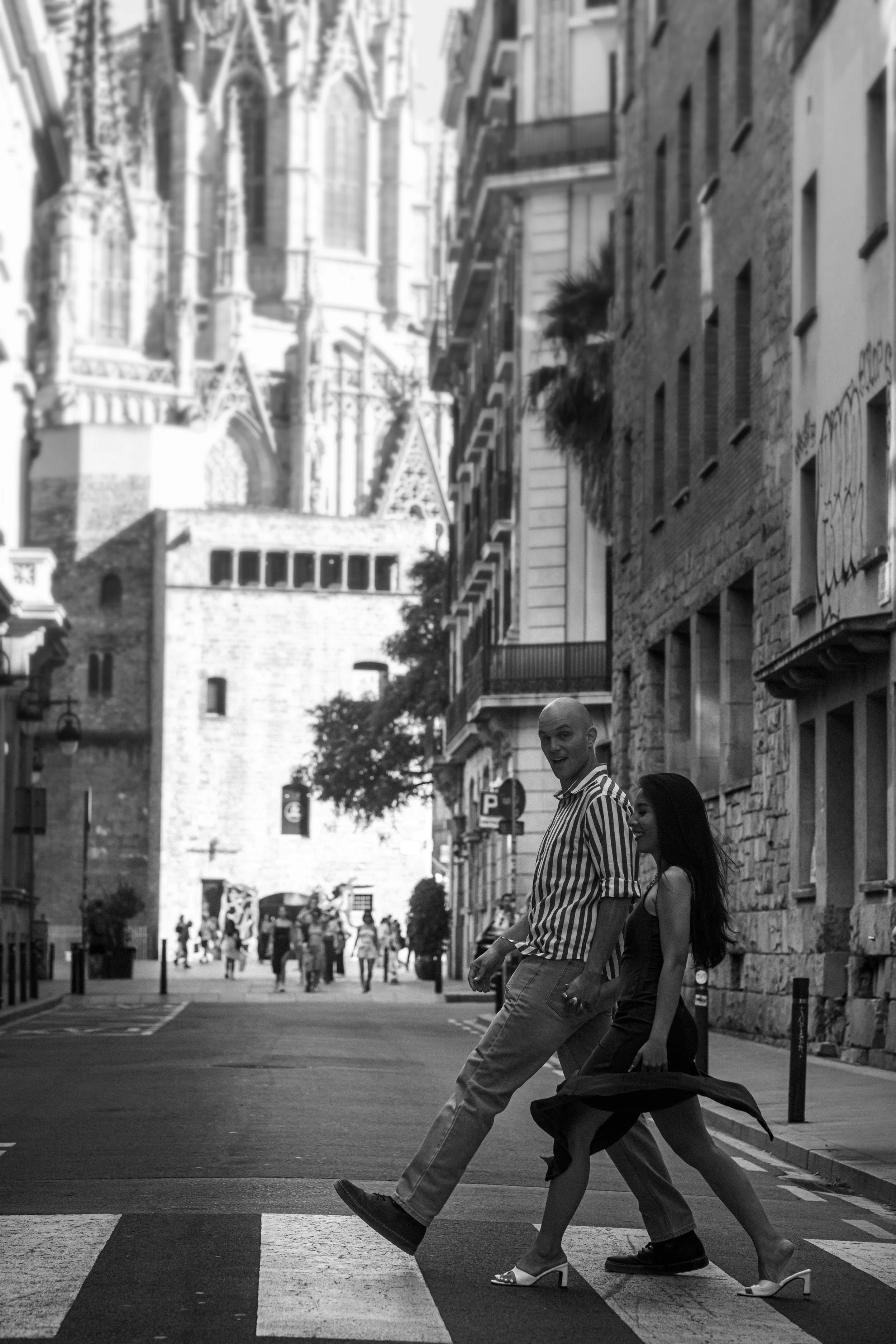 A black and white photo of a couple crossing a city street. The man is smiling, wearing a striped shirt and pants, with one foot lifted. The woman, wearing a dress and high heels, is smiling and holding a jacket. Tall buildings and a Gothic-style chu