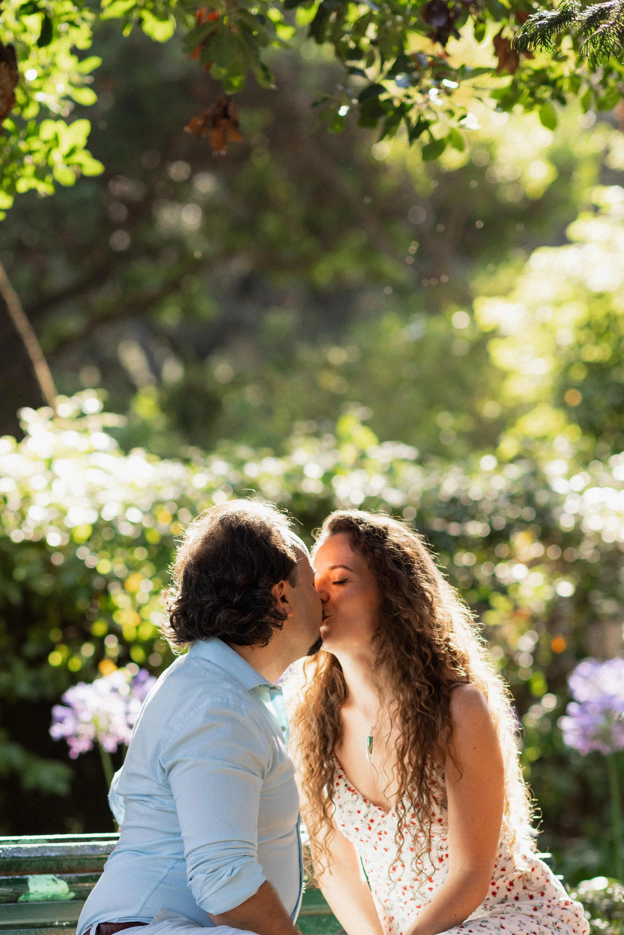 A couple sharing a kiss outdoors on a sunny day, surrounded by greenery and blooming flowers, sitting on a green park bench.