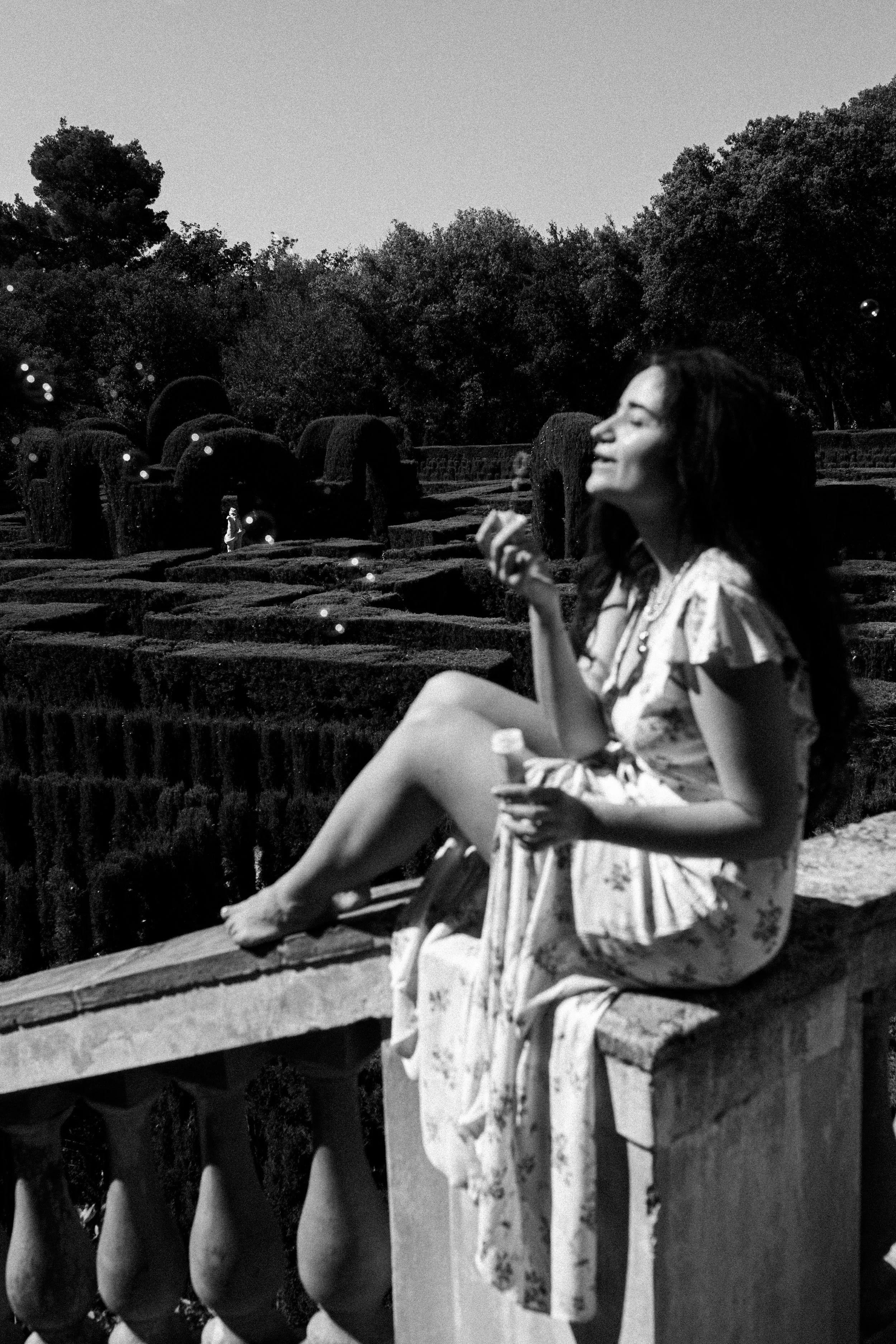 A woman in a floral dress sits on a stone railing outdoors, holding an ice cream cone, smiling with her eyes closed. In the background, there are manicured bushes and trees, and a small figure walking in the distance.