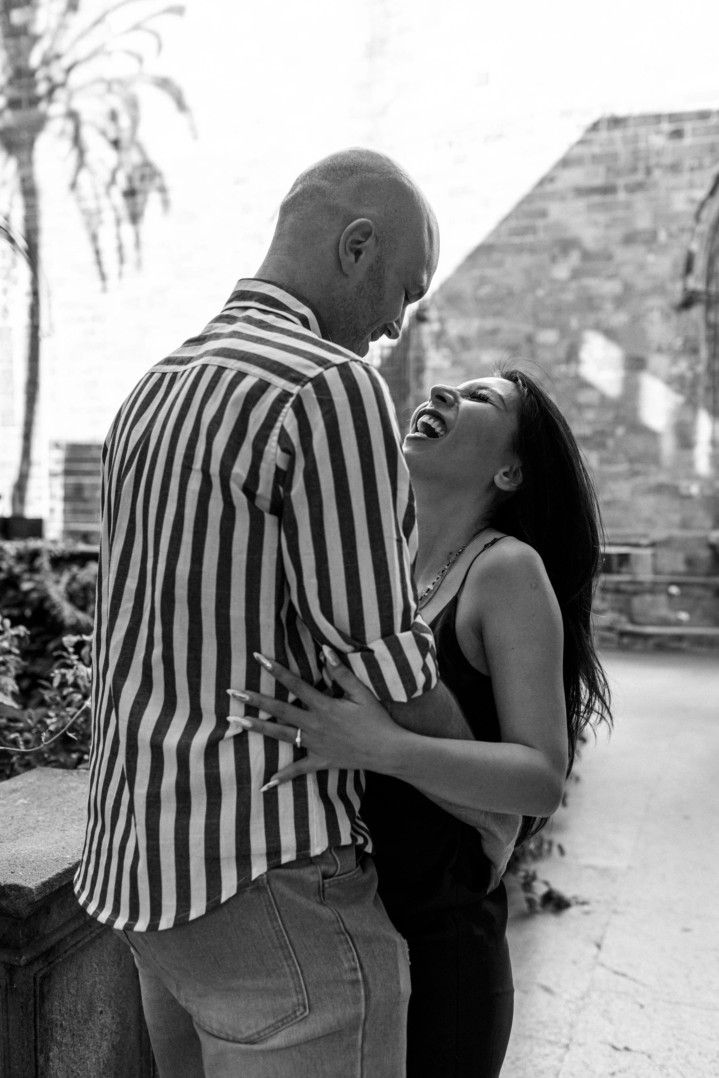 A black-and-white photo of a man and woman laughing and embracing outside, with a brick wall and urban background.