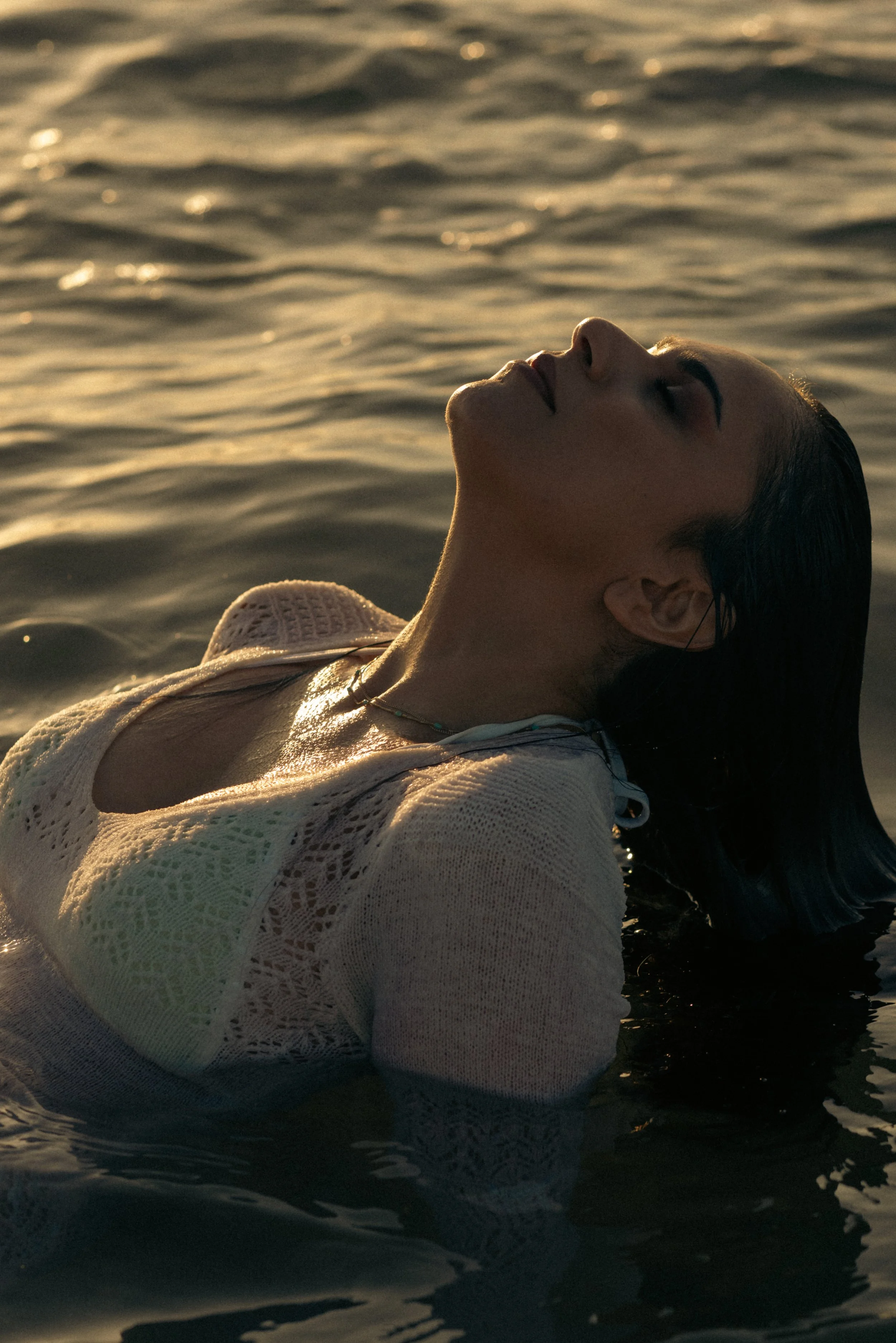 A woman is floating in water during sunset with her eyes closed, wearing a white lace dress and a necklace.