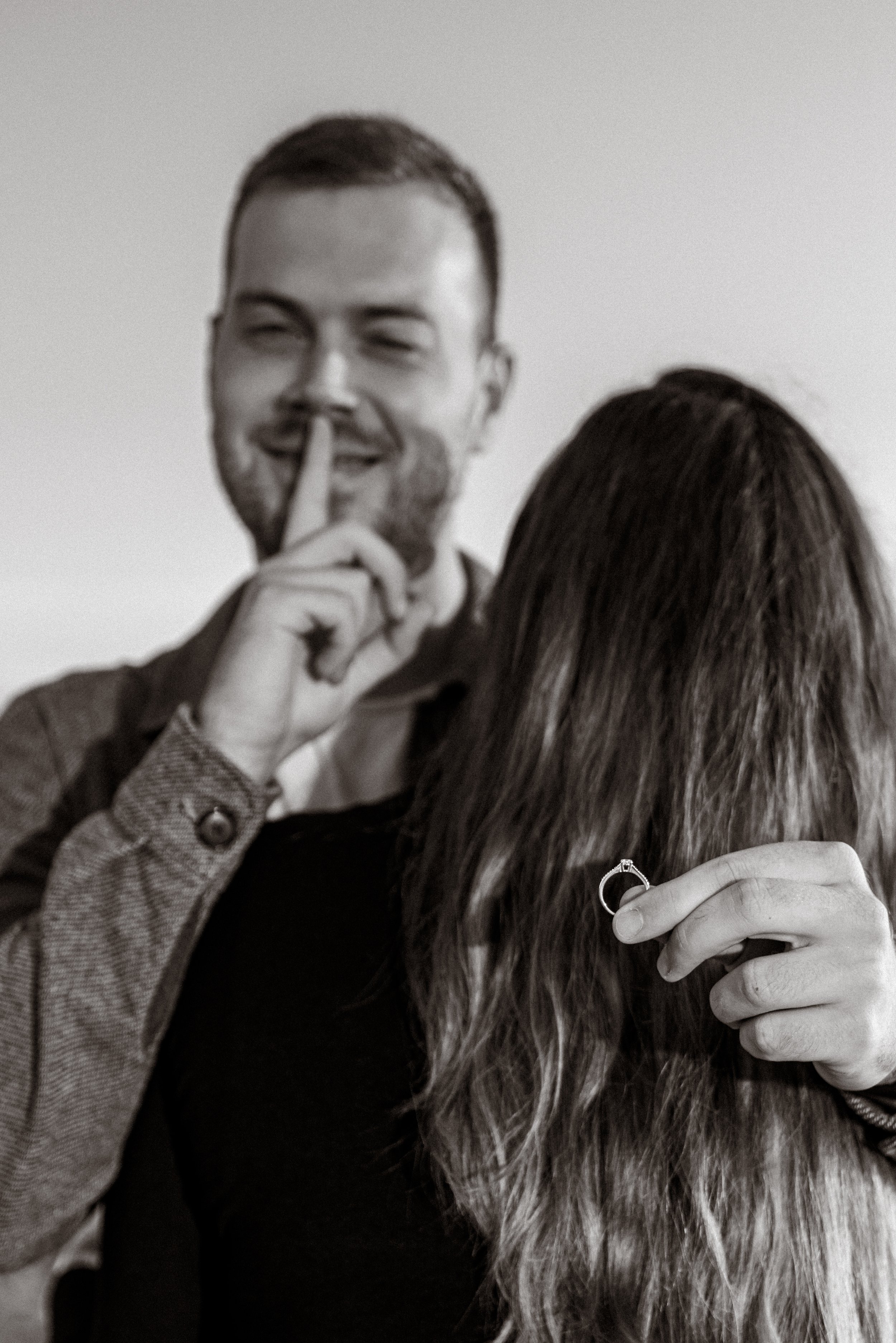 A man holding a ring, secretly placing a finger on his lips to indicate silence, while standing behind a woman holding a ring. The woman has long hair covering her face, and the man has a mischievous smile.