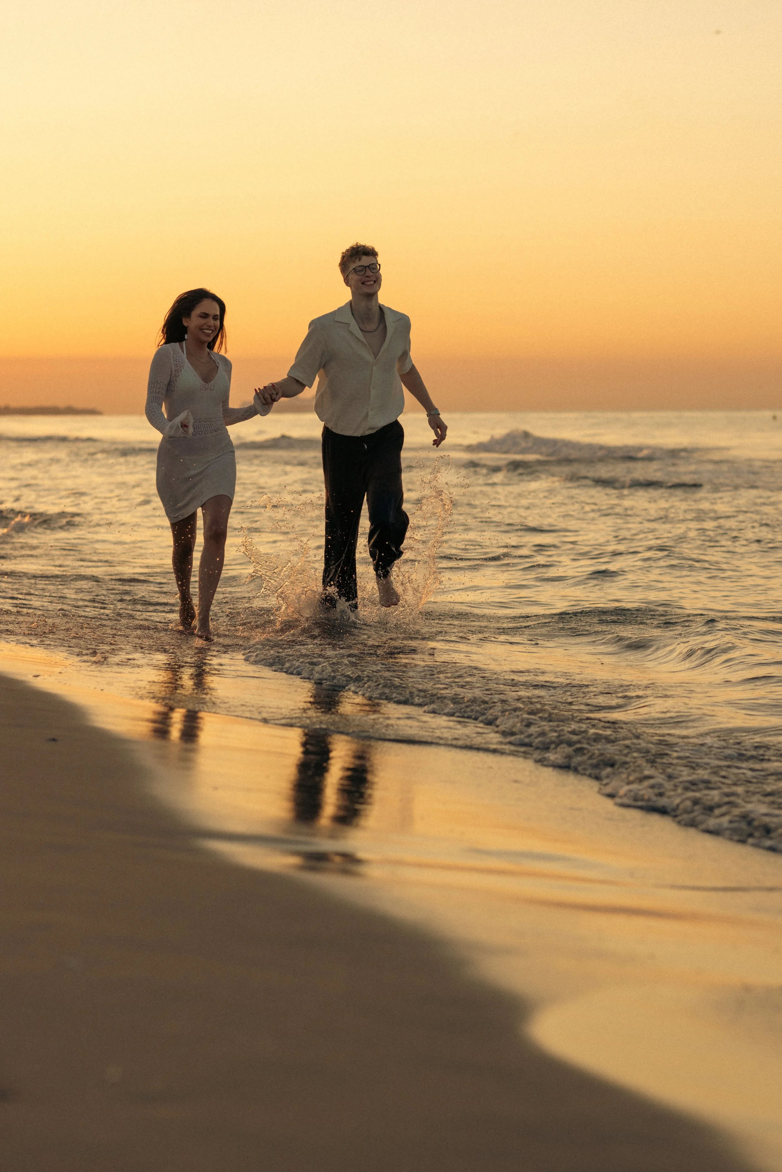 A man and woman holding hands, running along the shoreline at sunset, with the sky painted in warm hues.