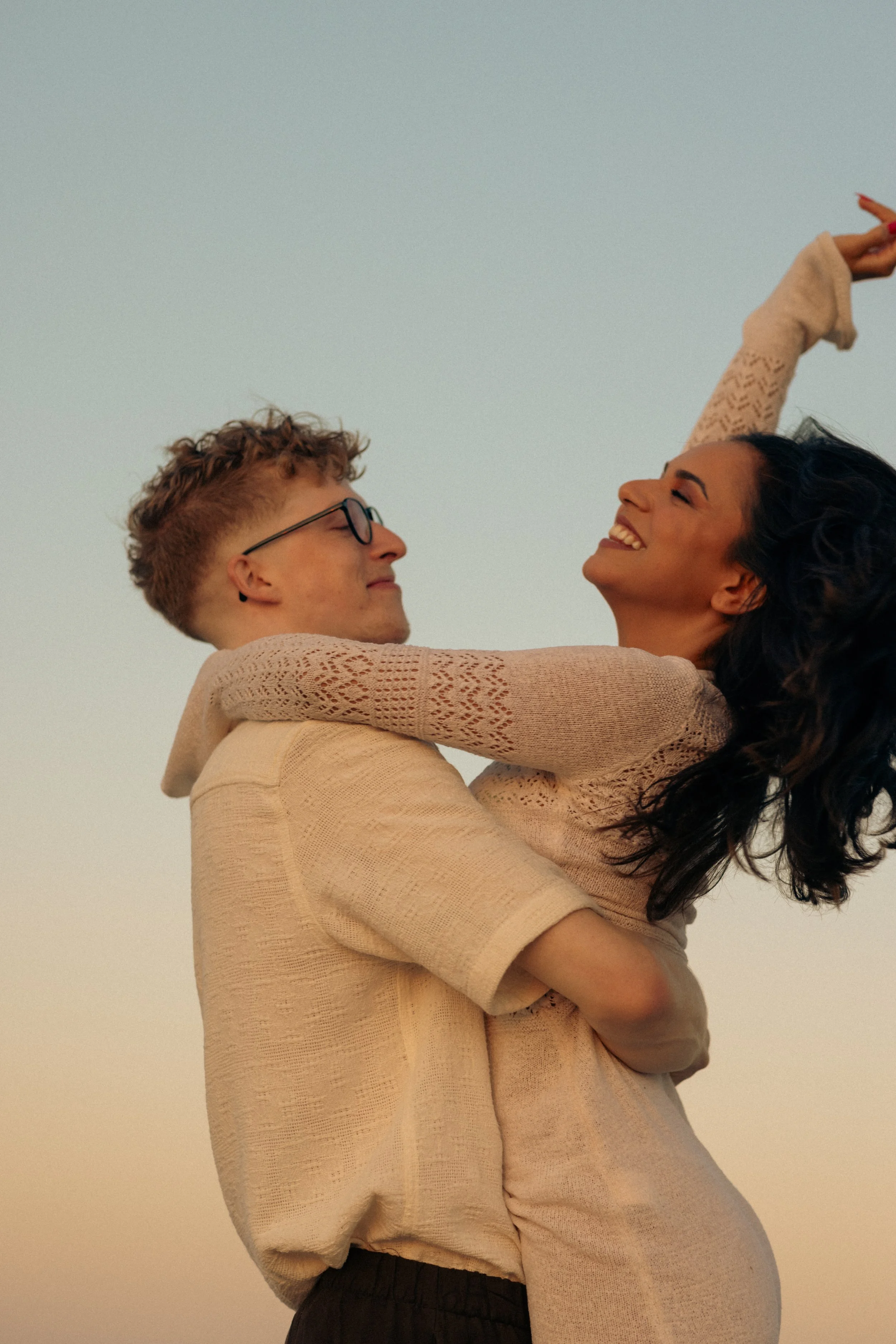 A happy couple, a man and a woman, enjoying a moment together outdoors at sunset, with the woman being lifted by the man. They are smiling and appear to be very joyful.