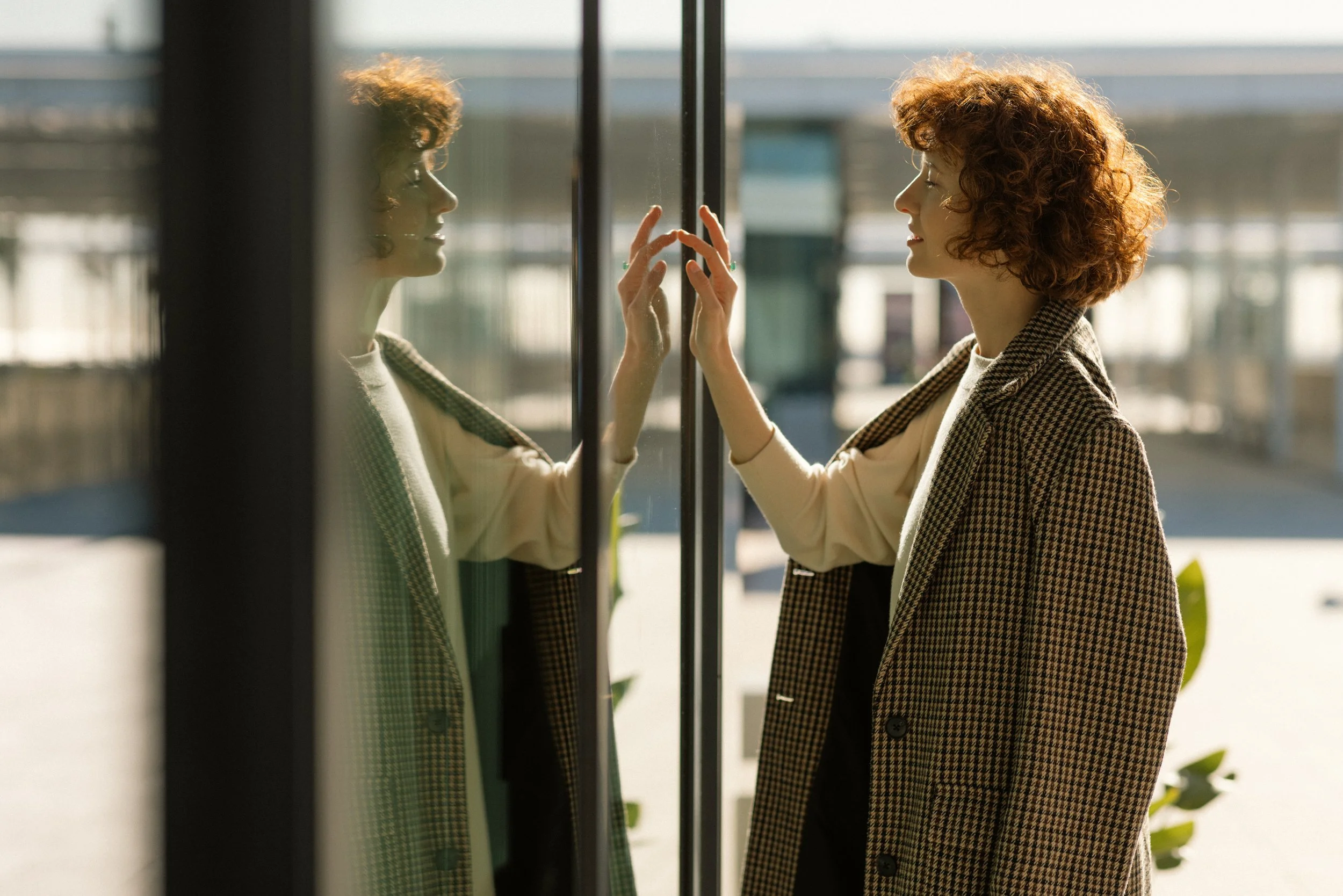 A woman with curly red hair and a checked blazer touches a glass window, looking at her reflection outside.