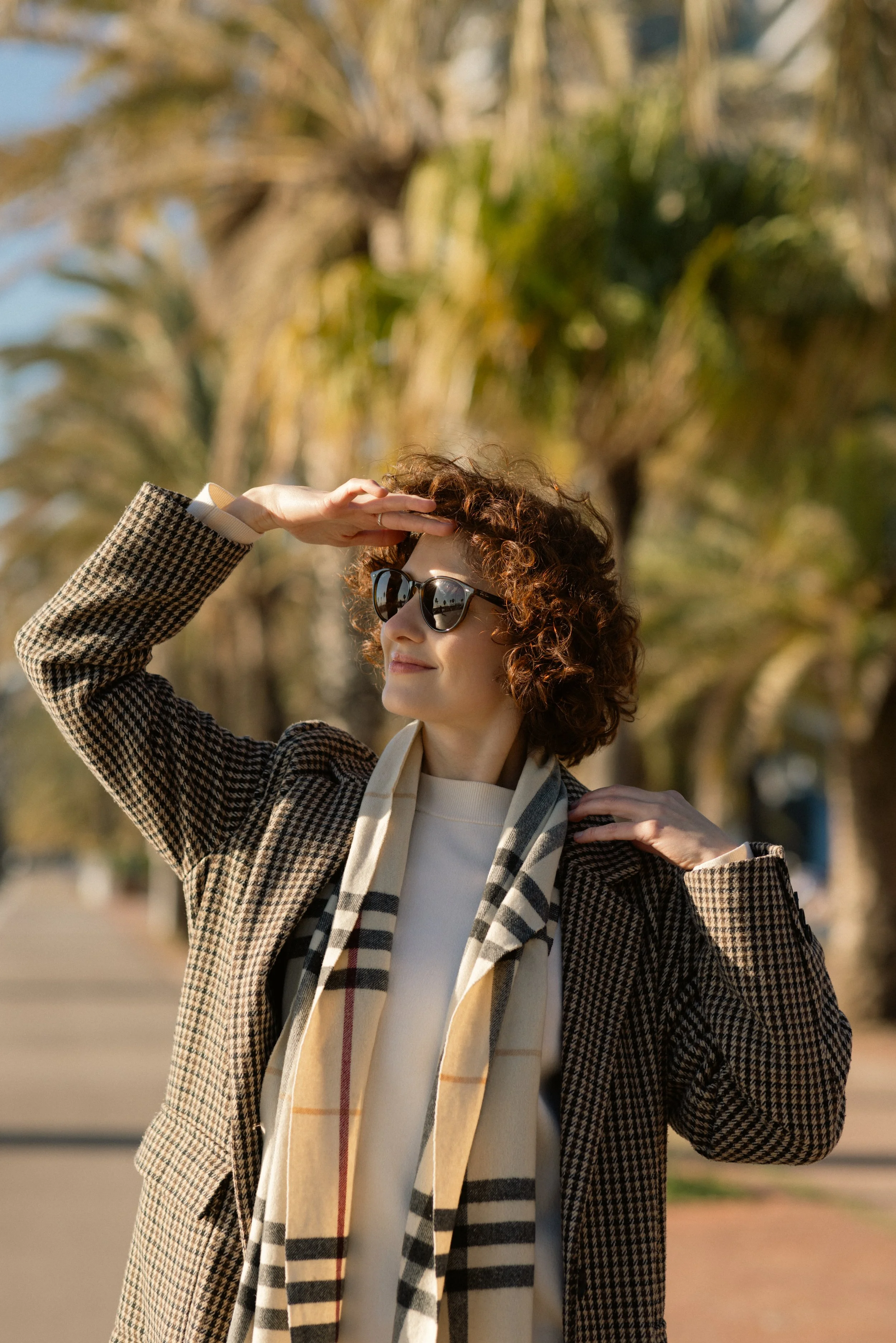 A woman with short curly hair and sunglasses, dressed in a checkered blazer and a beige scarf, standing outdoors in front of trees, with her hand shielding her eyes from the sun.