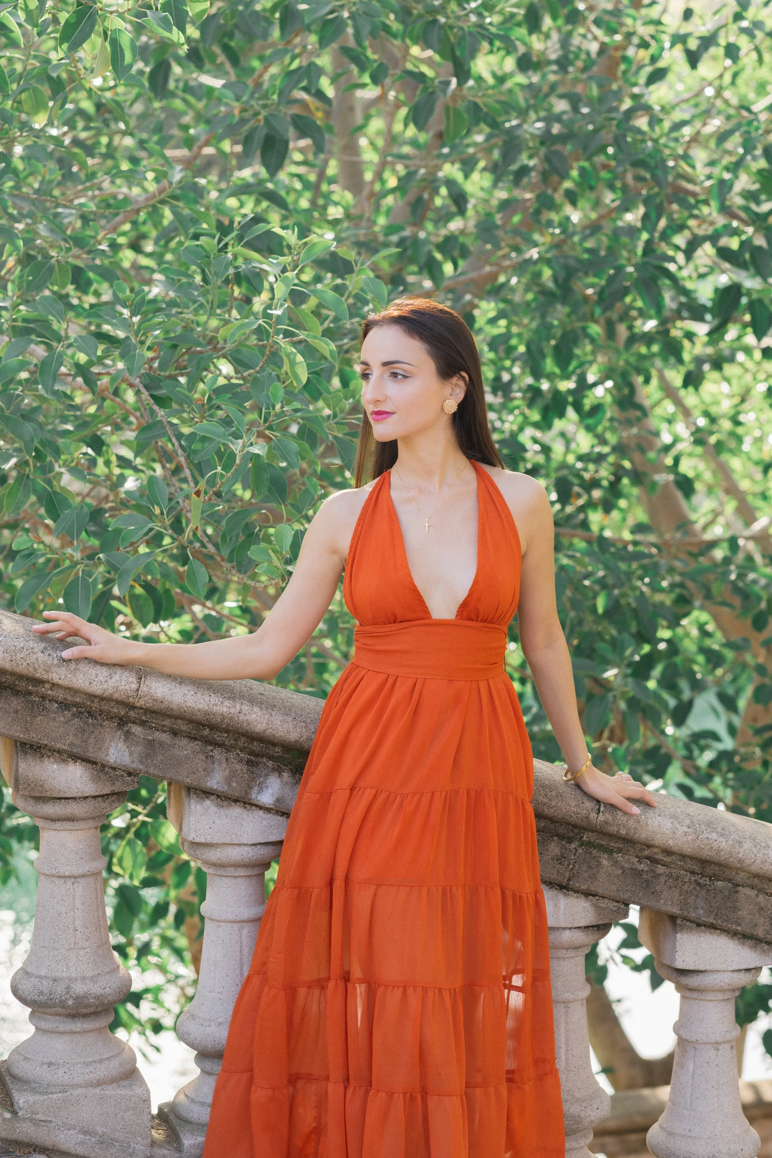 A woman in an orange dress leaning on a stone railing outdoors with green leafy trees in the background.