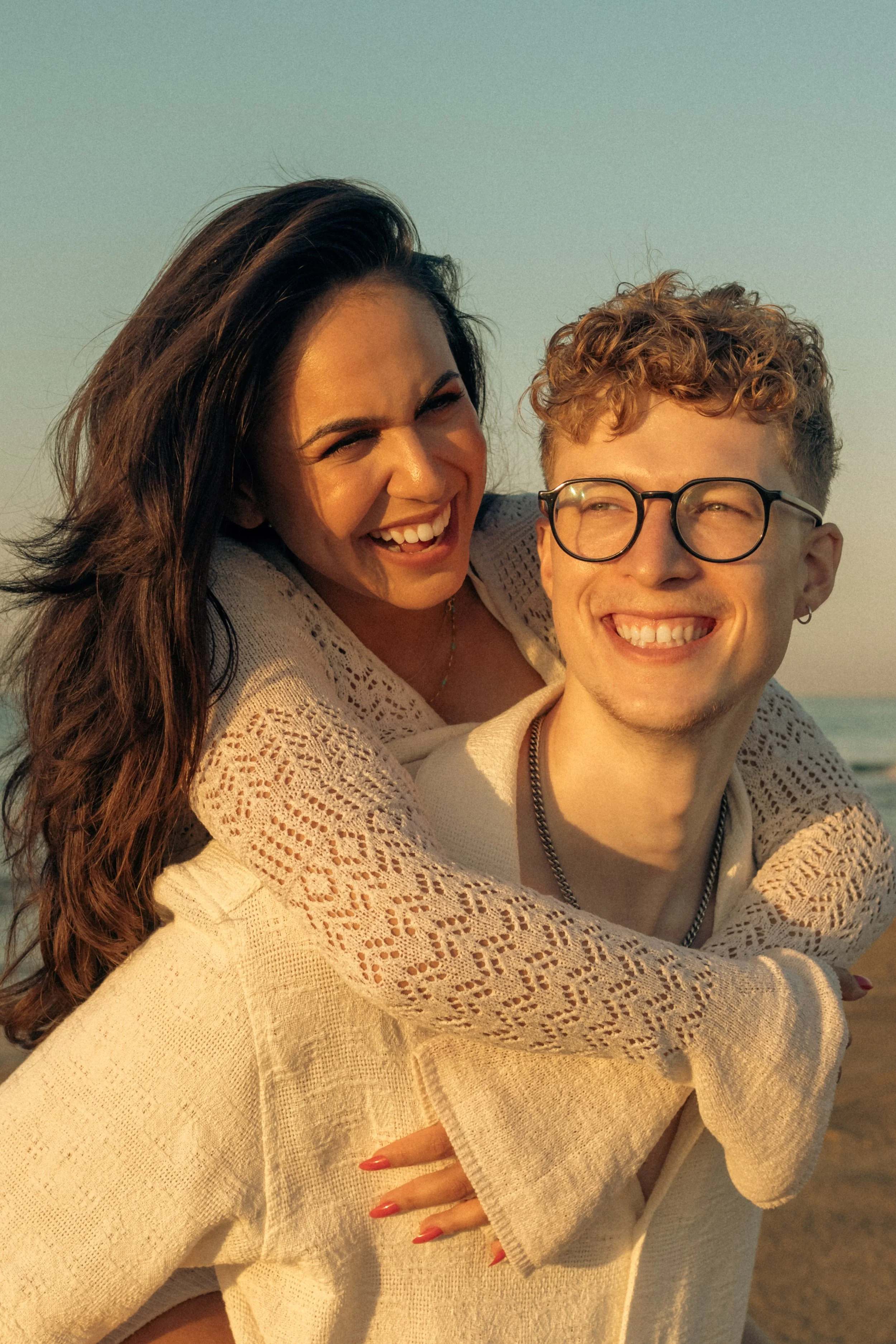 A smiling woman giving a piggyback ride to a smiling man on the beach during sunset.