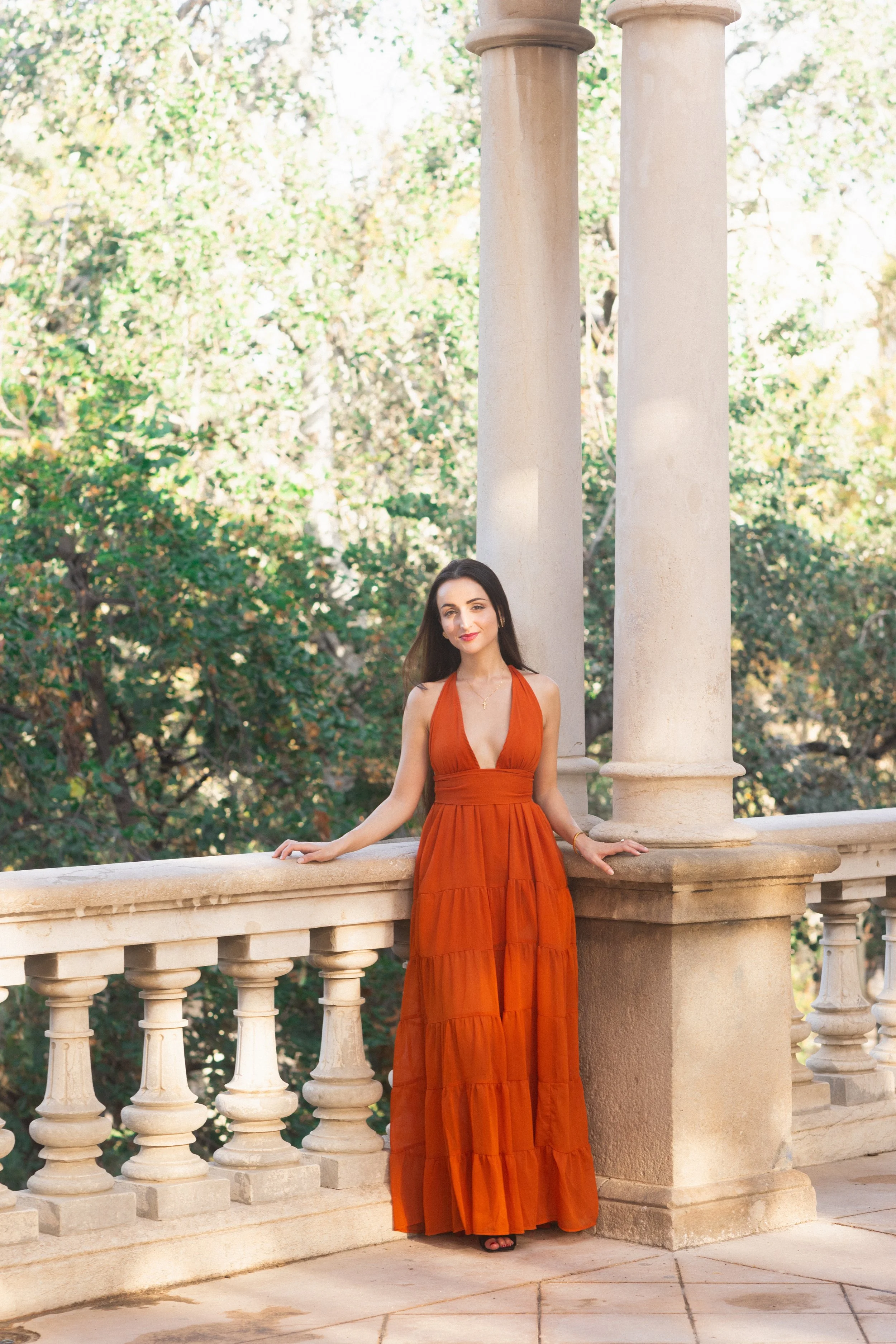 A woman in an orange maxi dress standing outdoors on a stone balcony with columns, with trees in the background.