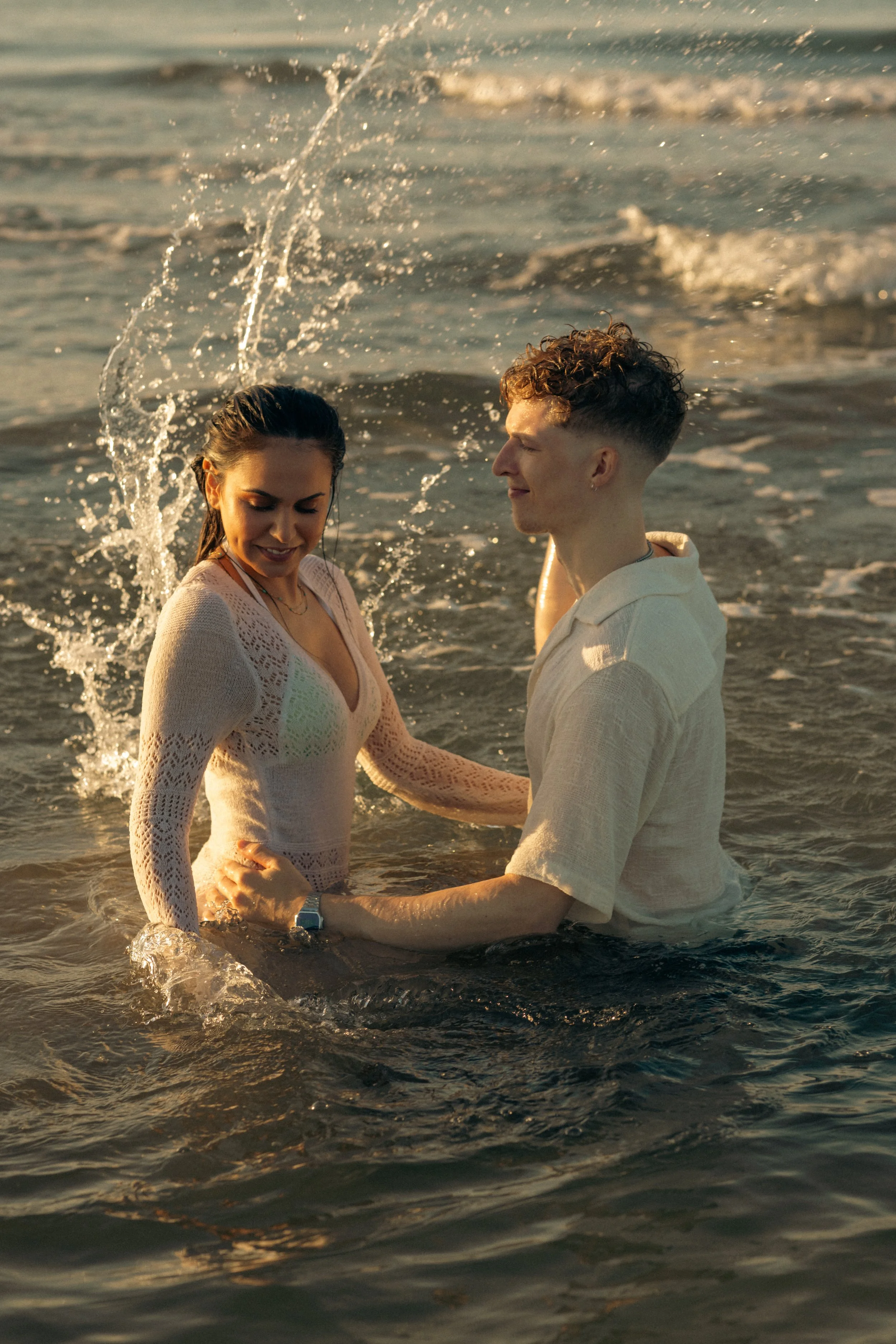 A young couple stands in the shallow water at the beach during sunset, holding hands and smiling for each other.