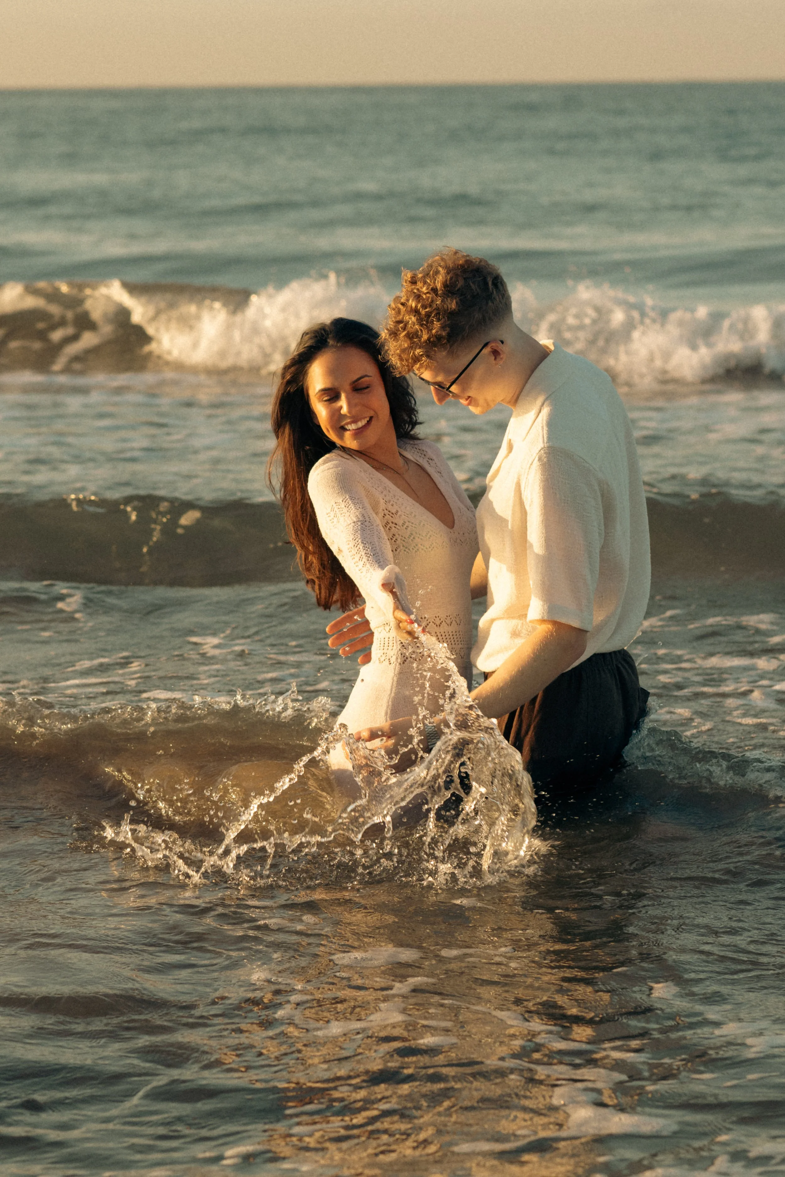 A couple standing in the shallow ocean water at sunset, smiling and holding hands, with waves in the background.