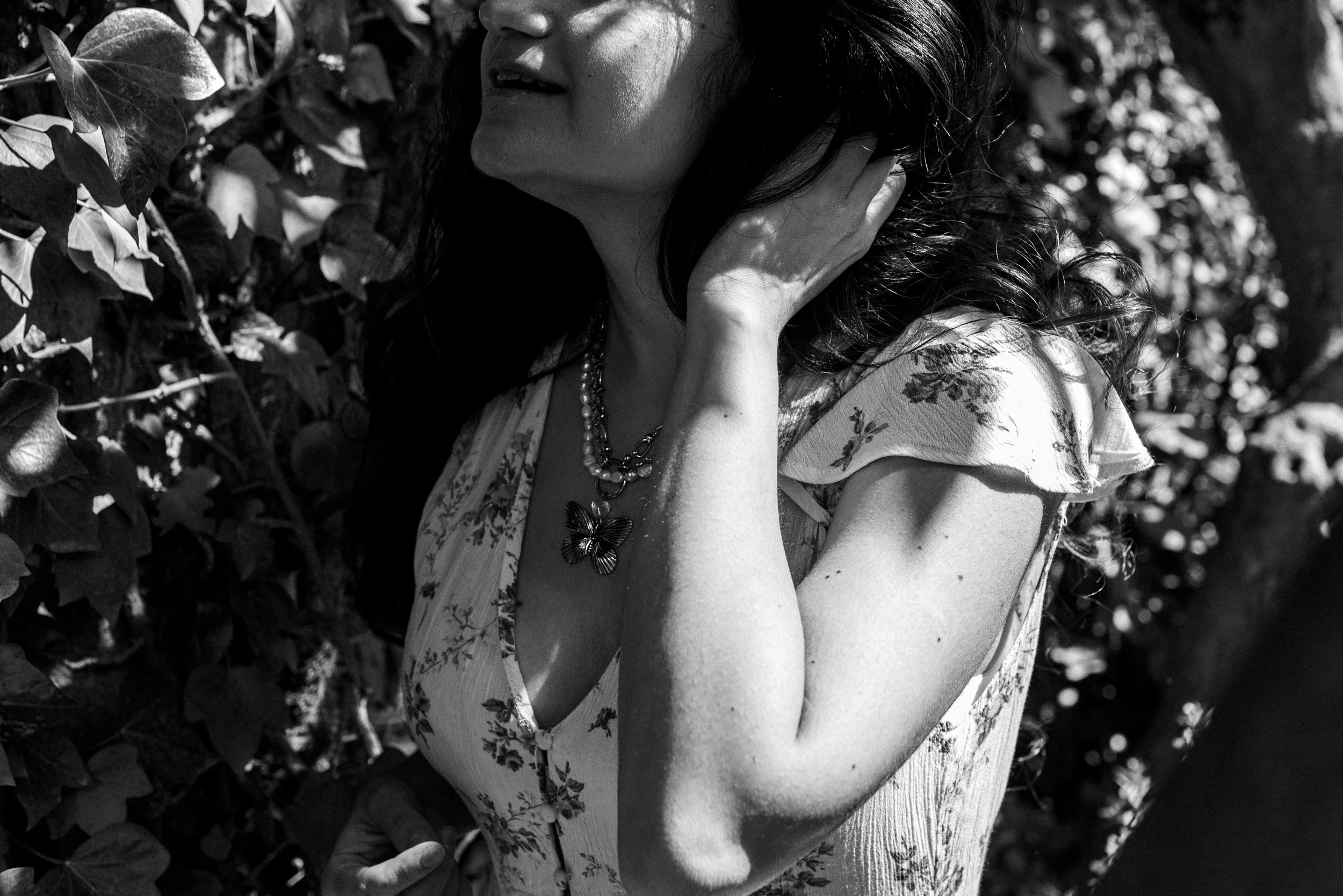 A woman with dark hair wearing a floral dress, a necklace with a butterfly pendant, standing among foliage.