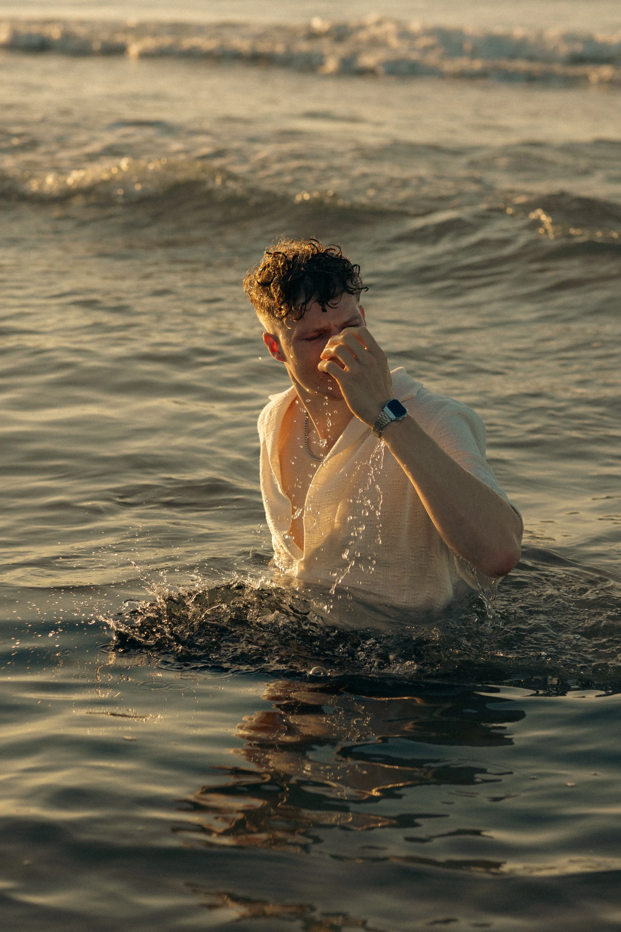 A young man with curly hair wearing a light-colored shirt and a watch is in the sea at sunset, covering his face with his hand.