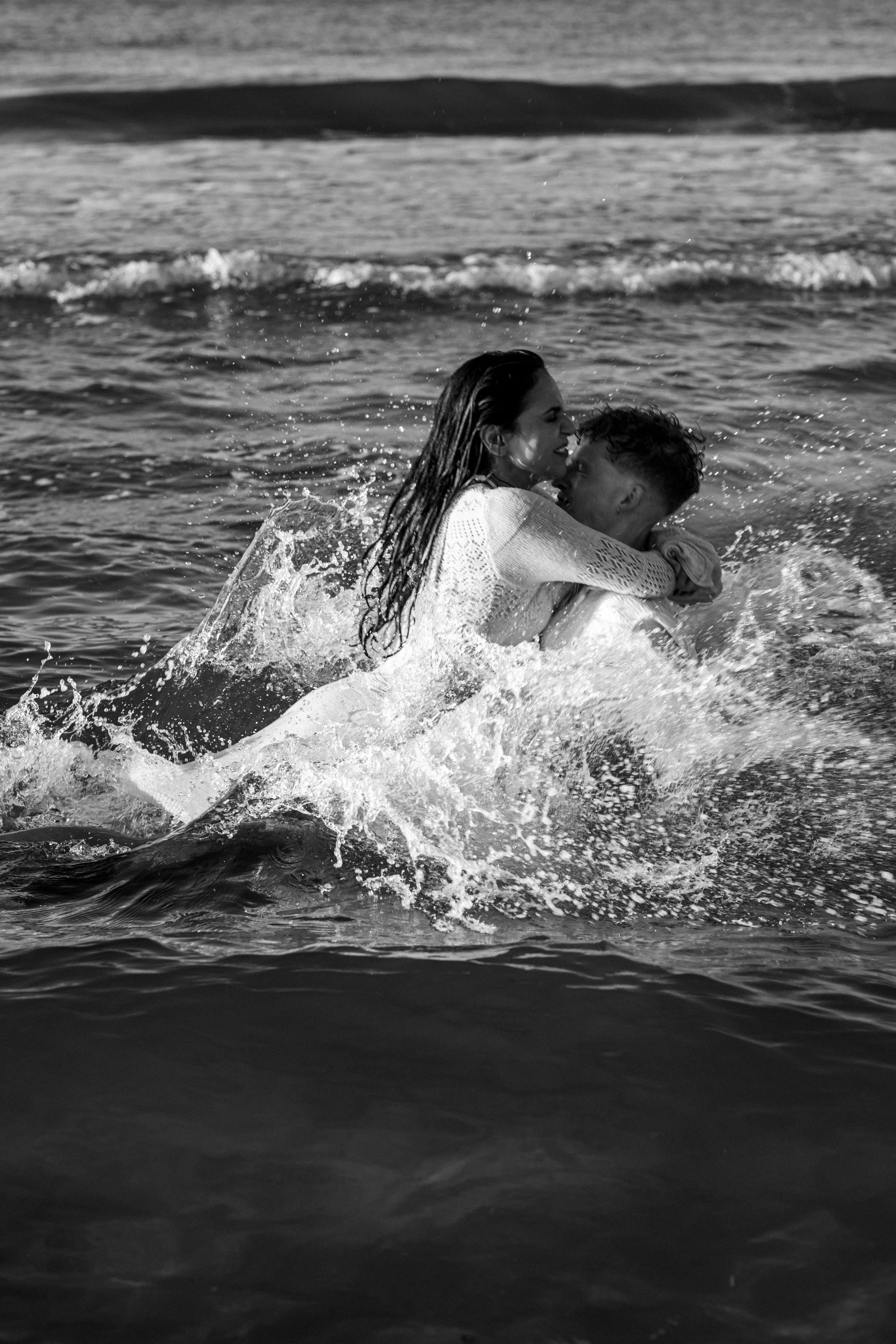 A black and white photo of a woman and a man in the ocean, the woman is holding onto the man, and they are surrounded by splashing water.