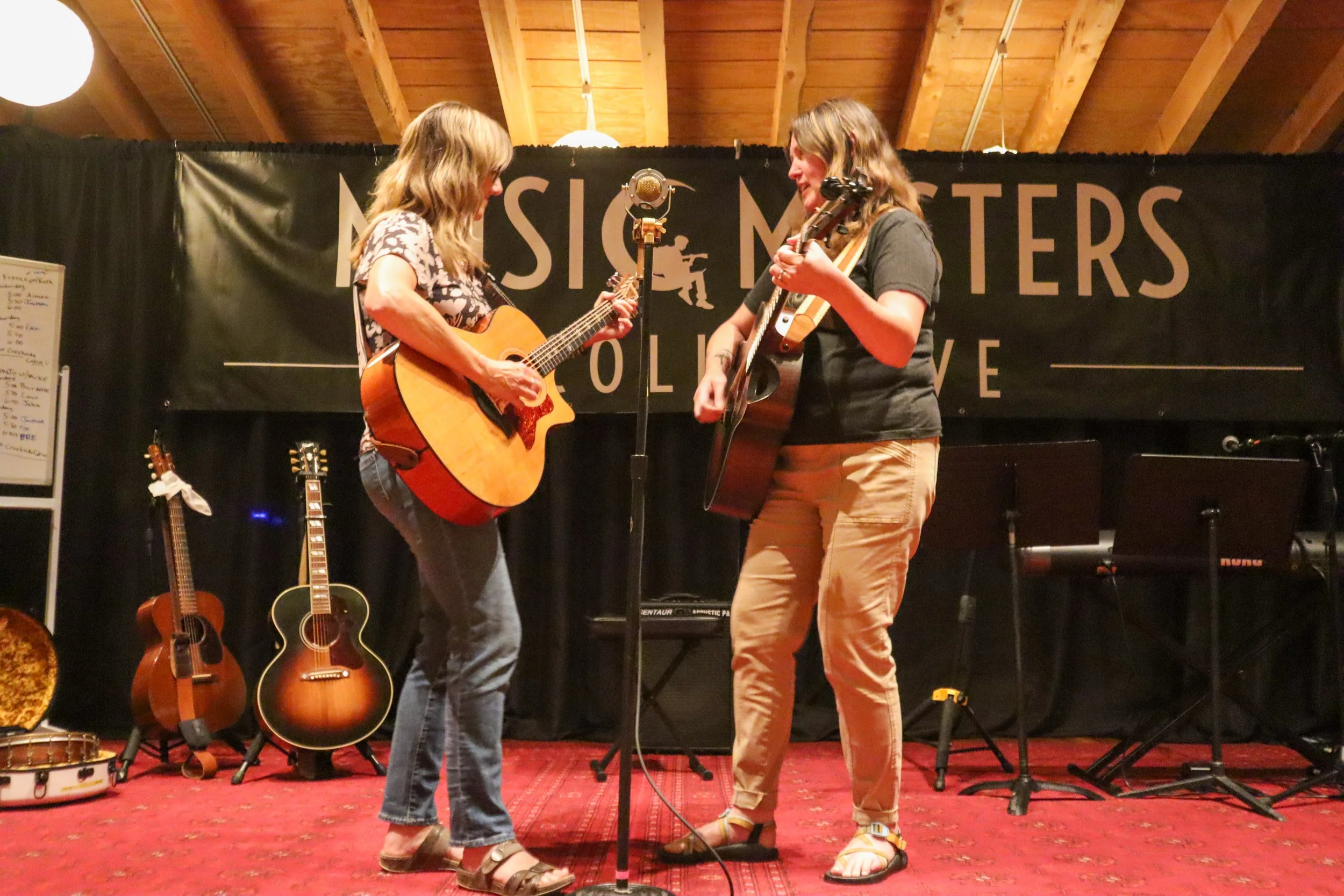 Two women playing acoustic guitars on stage during a live music performance, with a backdrop that reads "Music Masters". There are additional guitars and a keyboard on the stage.