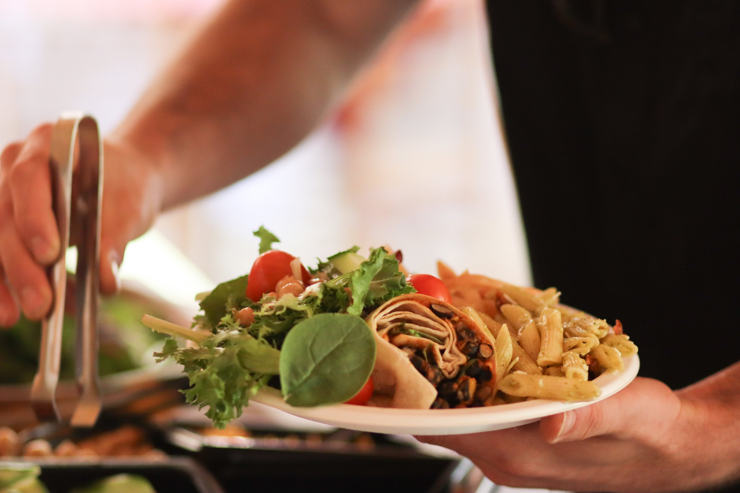 Person serving a plate of pasta, salad, and grilled vegetables at a buffet.