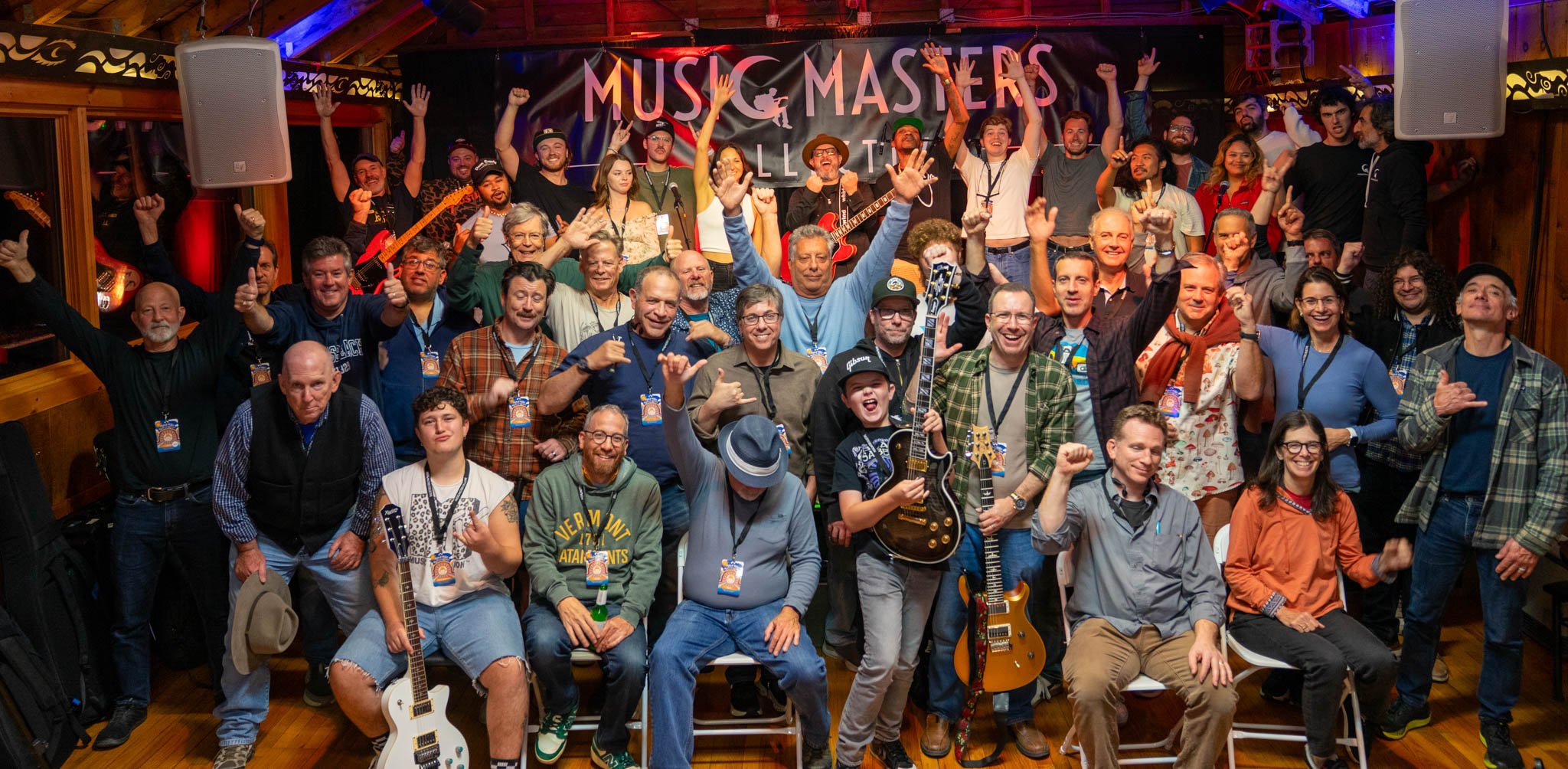Group of people at a music event called Music Masters, some holding guitars, celebrating and smiling, with a stage and musical instruments in the background.