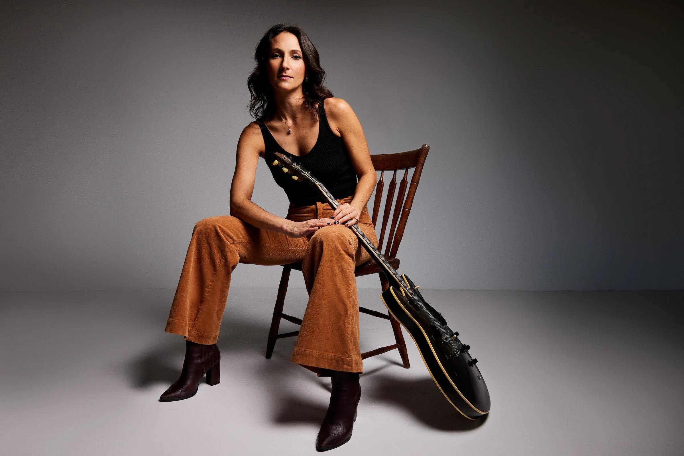 A woman with dark wavy hair sitting on a wooden chair holding a black electric guitar in her lap. She is wearing a black sleeveless top, brown wide-leg pants, and dark ankle boots. The background is plain and grey.